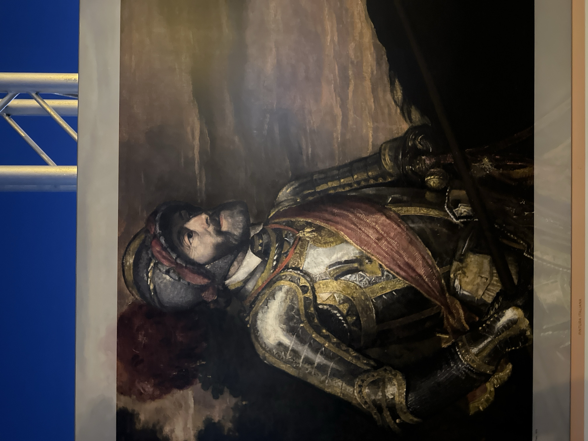 The image is a photograph of a painting displayed in a room. The painting depicts a man in ornate armor, wearing a helmet with a red plume. He has a beard and a red sash across his chest. The background of the painting is a cloudy, atmospheric landscape. Below the painting, a label reads "PINTURA ITALIANA." The room itself is dimly lit, with the top of the frame showing a blue sky and silver metal scaffolding. The overall impression is of an art exhibition. The location in Mazarrón, Spain is not discernible from the visual information provided.