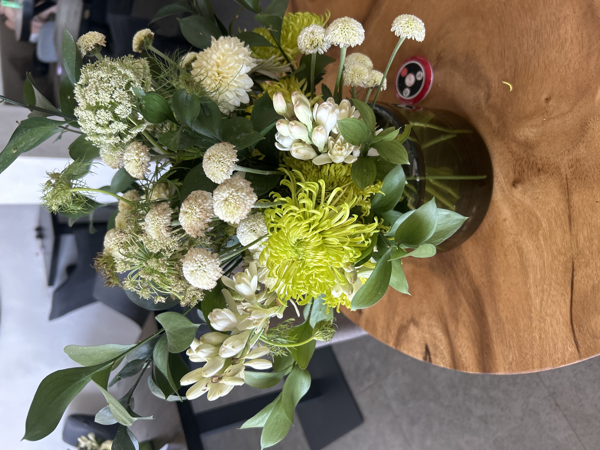 A high-angle shot captures a flower arrangement set on a wooden table, indoors. The arrangement features an array of white and green flowers, including white Queen Anne's Lace, white dahlias, white spherical flowers on thin stems, green spider mums, and white lilies, accented with green foliage. The vase holding the arrangement is made of dark glass. Adjacent to the vase on the wooden table, a red and black object with buttons is also visible. The image is taken in Kuta Selatan, Indonesia.