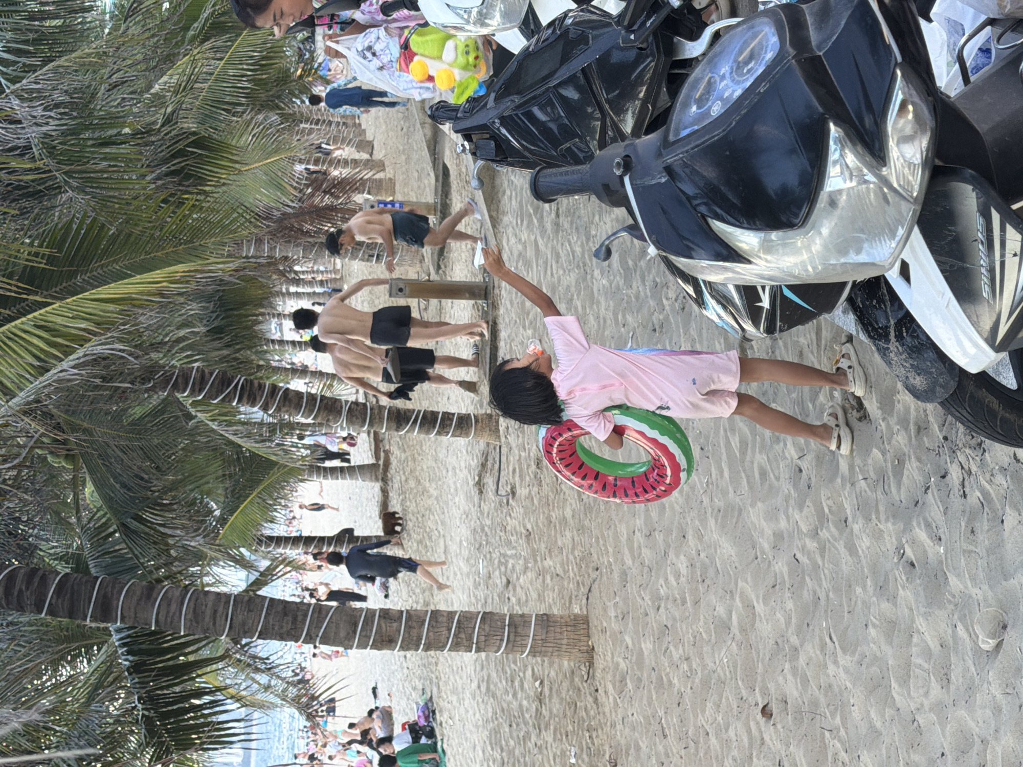 The image depicts a beach scene in Nha Trang Ward, Vietnam. The foreground features a young child wearing a pink shirt and white shoes, holding a watermelon-patterned inflatable ring. To the right of the child is a motorcycle. In the mid-ground, several people are visible on the sandy beach. Some are walking, while others appear to be engaged in activities or resting. Palm trees with white wrapping around their trunks are scattered across the scene, providing shade. The background shows the ocean and a crowded beach with many people, suggesting it's a popular spot. The overall impression is a lively, sunny day at the beach, likely during the day given the bright lighting. There are no discernible emotions being conveyed. No visible text is present in the image.