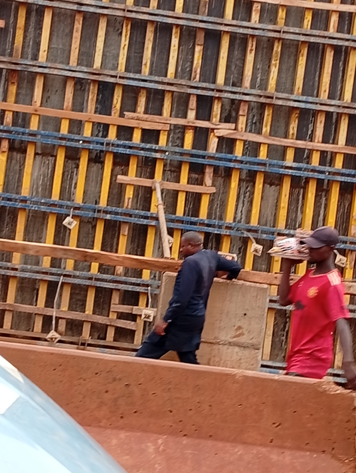 The image shows two men on a construction site in Katambe, Nigeria. The background is dominated by a wooden and metal formwork structure, likely for concrete pouring. The formwork consists of vertical wooden planks painted yellow and dark gray, interspersed with horizontal blue metal bars. Some wooden planks are also visible within the structure.
The subject on the right is a man wearing a red t-shirt with a crest and a black baseball cap. He is carrying a flat object, possibly a piece of wood or material, on his head. The subject on the left is wearing a dark blue long-sleeved shirt and dark pants. He is leaning on a concrete block, looking away from the camera.
The scene suggests ongoing construction work. The lighting indicates it is daytime. The overall atmosphere appears to be one of work and labor. There is no visible text in the image.
