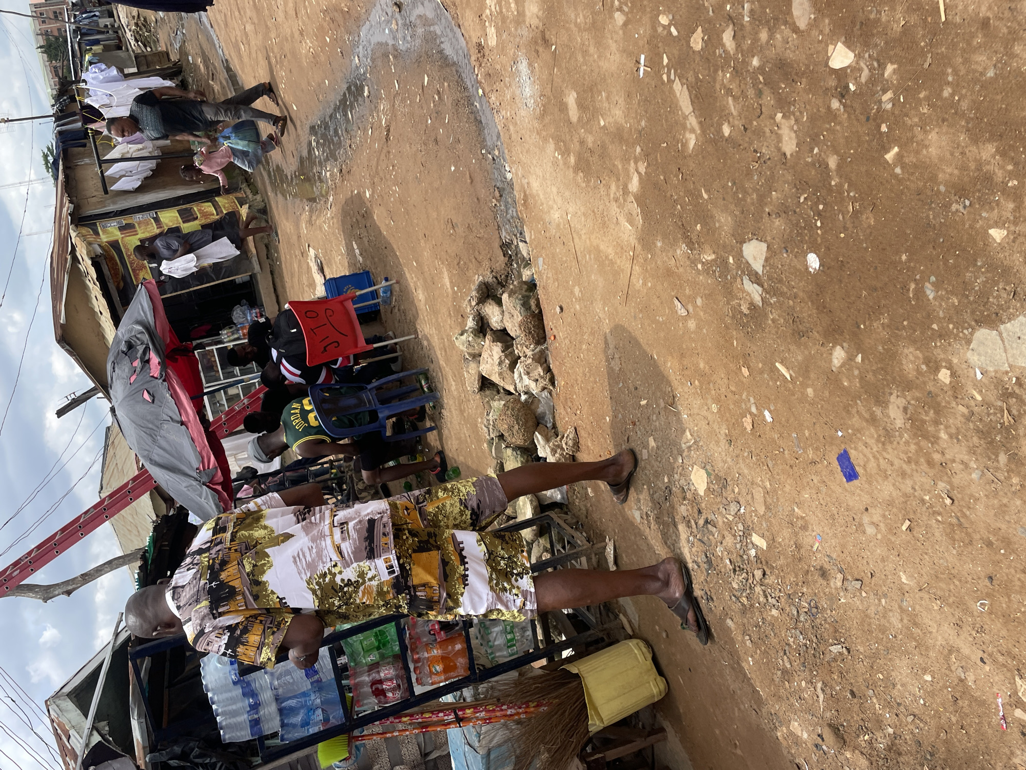 The image depicts a street scene in Abuja, Nigeria, characterized by a mix of informal structures and a bustling environment. In the foreground, a man wearing a brightly colored shirt and patterned shorts stands facing away from the camera, appearing to be browsing at a stall. To his left, a rack displays several bottles of beverages, including water and other drinks. A yellow bucket and a bundle of brooms are also visible near the stall.
In the background, several makeshift shops are lined up, constructed from corrugated iron and wood. Clothes are hung out to dry on lines strung between the structures. A group of people are gathered around tables and chairs, some appearing to be engaged in conversation. One person is walking away from the viewer, holding a blue bag, and is accompanied by a young girl. The ground is uneven and dusty, with scattered debris. A small stream of water runs through the scene. The sky is overcast with clouds. The overall impression is of a lively, everyday scene in a developing urban area.