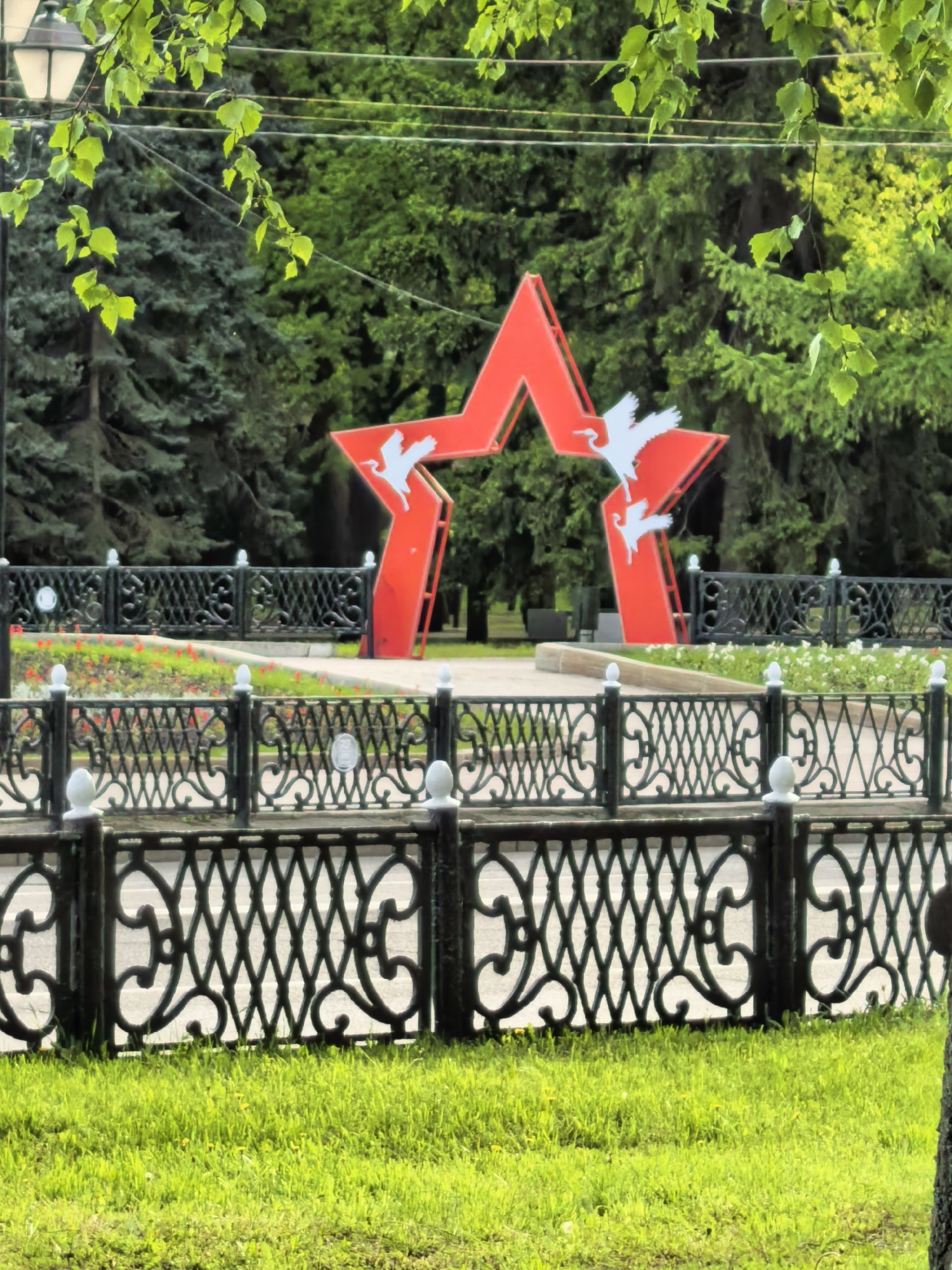 The image shows a monument in the city of Sterlitamak, Russia. The monument is a large red star with white cranes flying through it. It is surrounded by a decorative black metal fence. There are green trees in the background and a well-maintained lawn in the foreground. A few flowering plants are visible inside the fence. The weather appears to be bright, and the scene is viewed from ground level through some foliage.