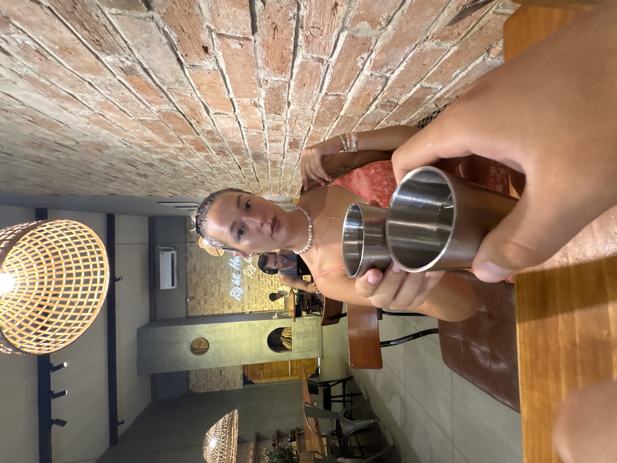 The image depicts a close-up view of two hands holding metal shot glasses, engaged in a toast. In the background, a woman with her hair pulled back and wearing a coral-colored dress and a pearl necklace is seated at a table, looking towards the camera. She is adorned with bracelets. Behind her, the interior of a restaurant is visible, characterized by exposed brick walls, wooden furniture, and rattan pendant lights. The restaurant appears to be in Da Nang, Vietnam, as indicated by the neon sign in the background that reads "Right Here". Other patrons are seated at tables, and a man wearing headphones is working on a laptop in the middle ground. The overall atmosphere suggests a casual dining or bar setting.
