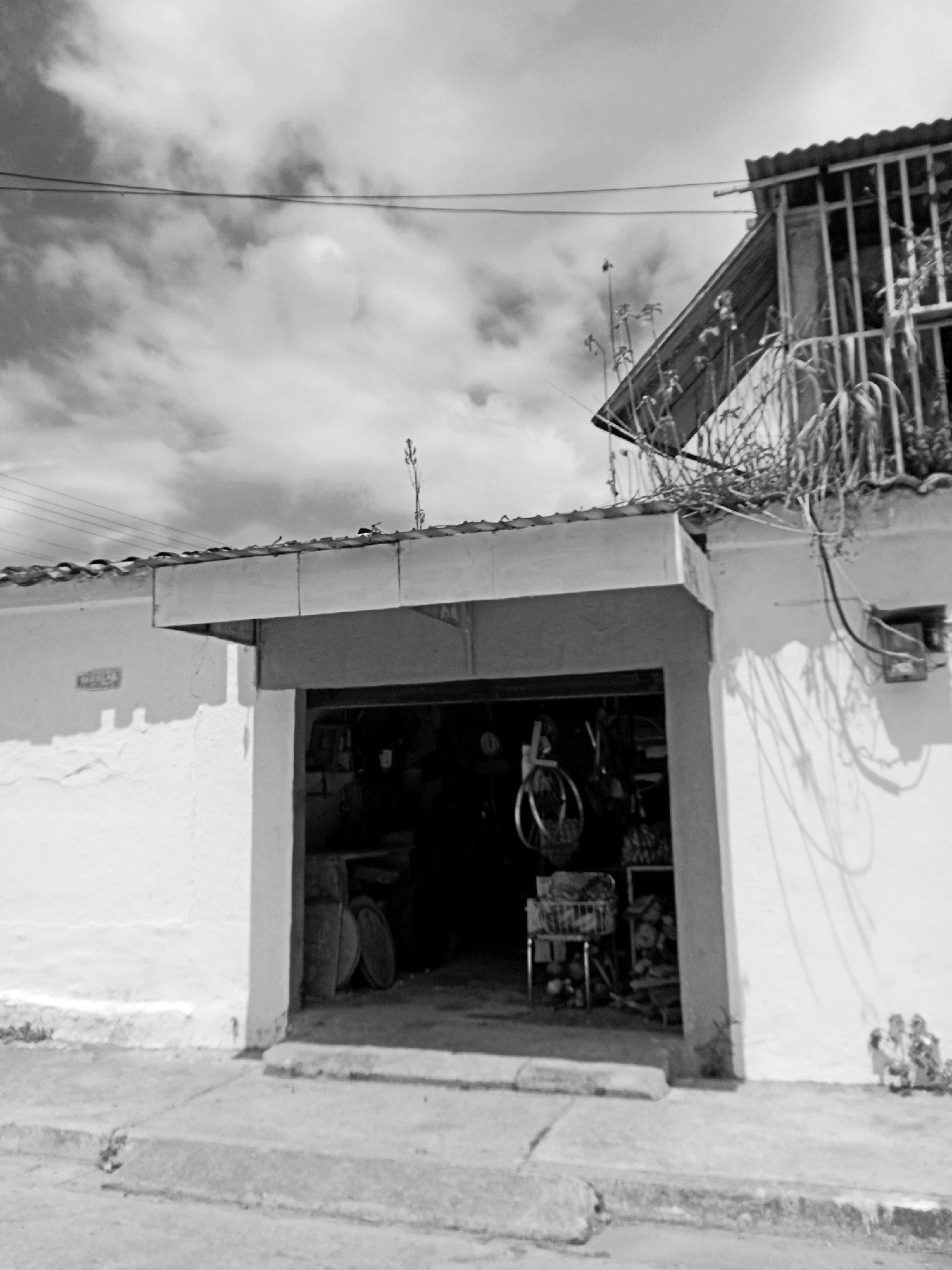 This black and white image depicts a street scene in Montalbán, Venezuela, focusing on the entrance of a building. The building has a white, weathered exterior with a partially open doorway. Inside the doorway, objects are vaguely discernible, including what appears to be a bicycle wheel, shelving, and miscellaneous items. The roofline of the building extends over the doorway, providing partial shade. Above the main building, another structure is visible with metal bars covering a window. Overhead, power lines stretch across a cloudy sky. A sidewalk runs in front of the building, and a small, unidentifiable object sits near the building's right corner.