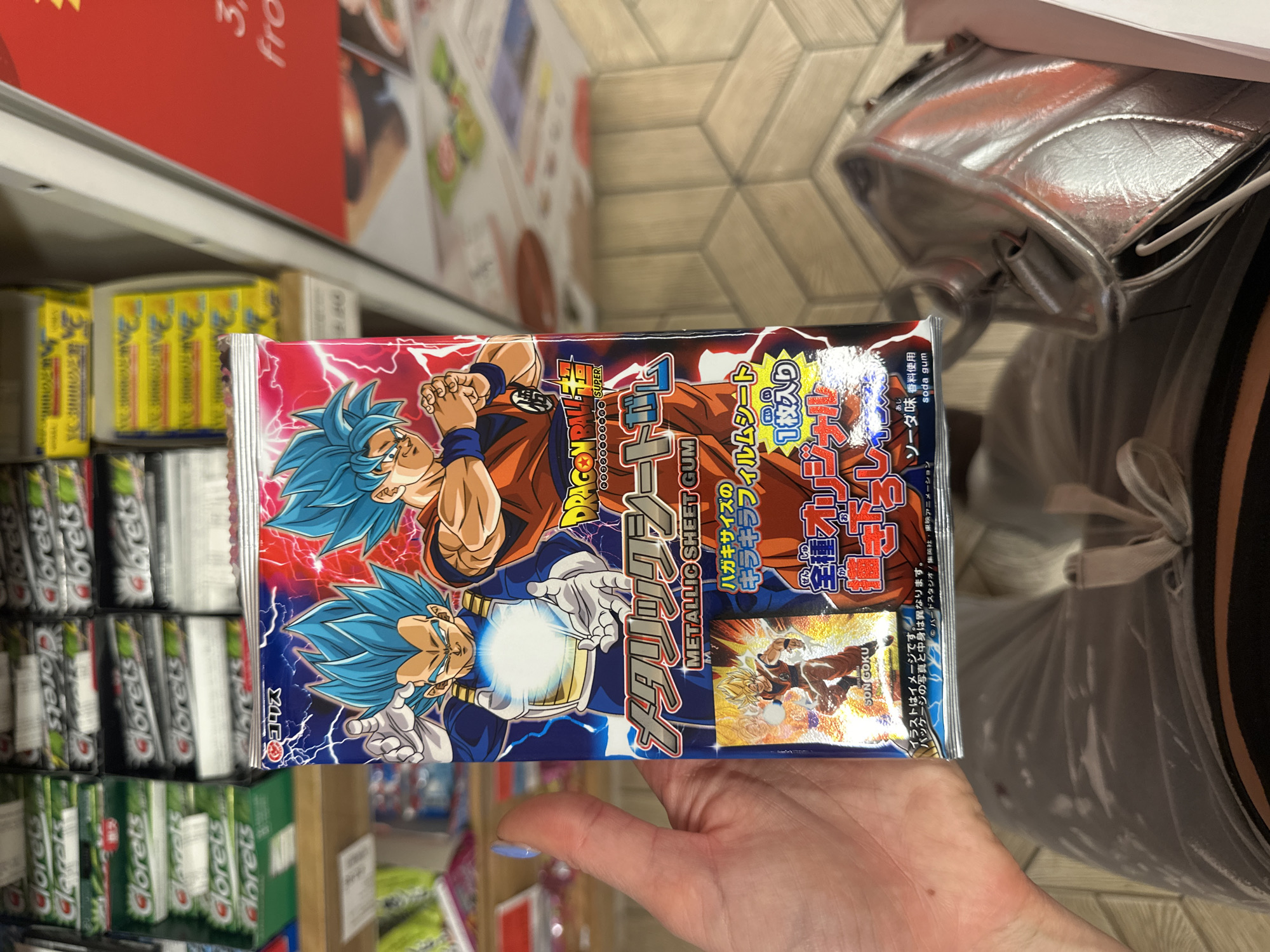 A person holds a package of Dragon Ball Super metallic sheet gum, featuring images of Goku and Vegeta, in a store aisle. Clorets gum packs are visible on a shelf to the left. A silver bag is to the right of the hand. The package's text is mostly Japanese. The location is likely a retail setting, but the city or country is not specified within the visual details of the image.