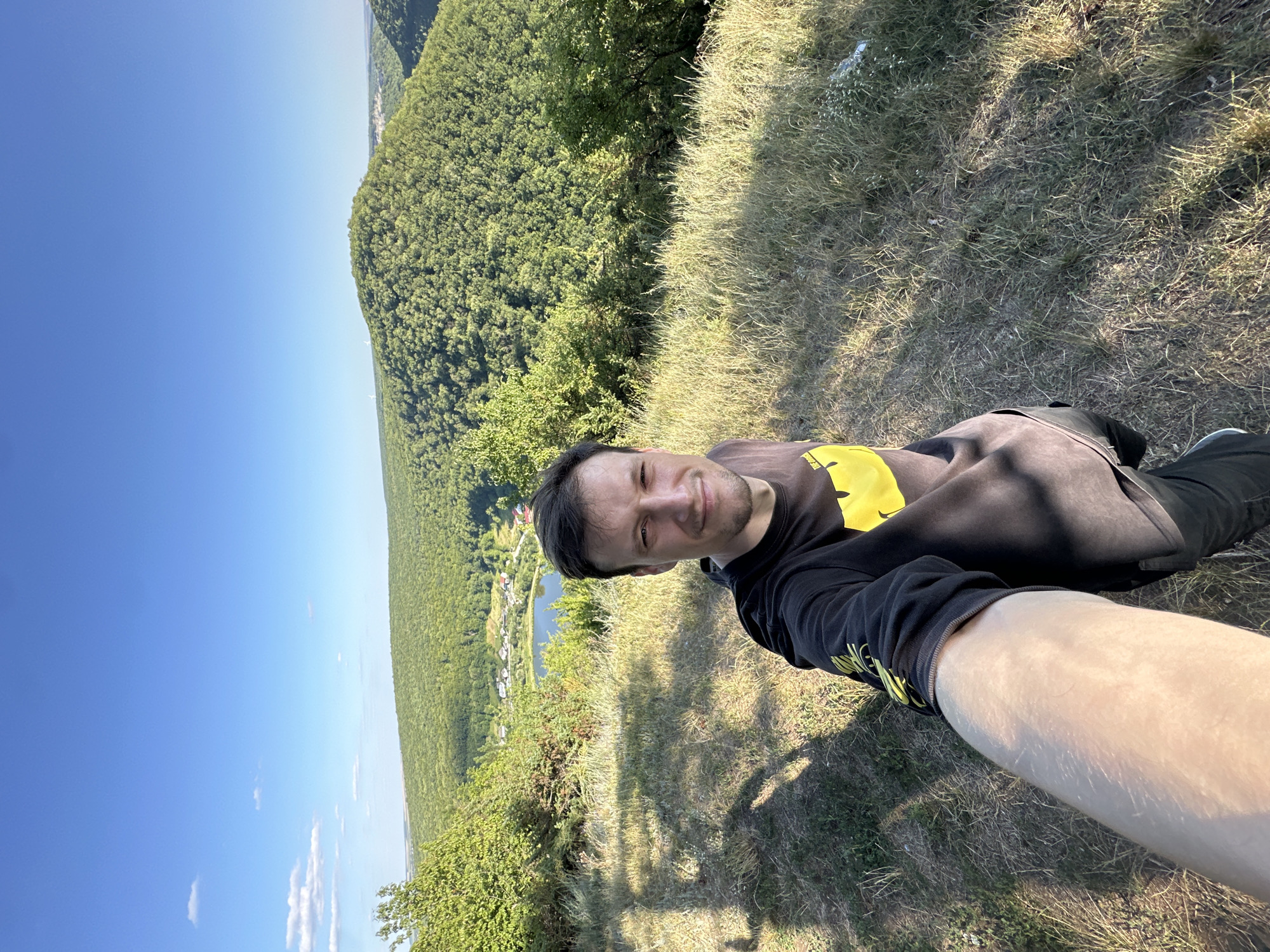 A male individual is taking a selfie on a grassy hill overlooking a valley in Pryvorottia Druhe, Ukraine. The man is wearing a dark long-sleeved shirt with a yellow graphic on it. He is smiling and looking directly at the camera. The background shows a lush green valley with a body of water, and rolling hills covered in trees under a clear blue sky. The sun is casting shadows on the ground, indicating it is likely late afternoon. The terrain appears natural and undeveloped, suggesting a rural or park-like setting.