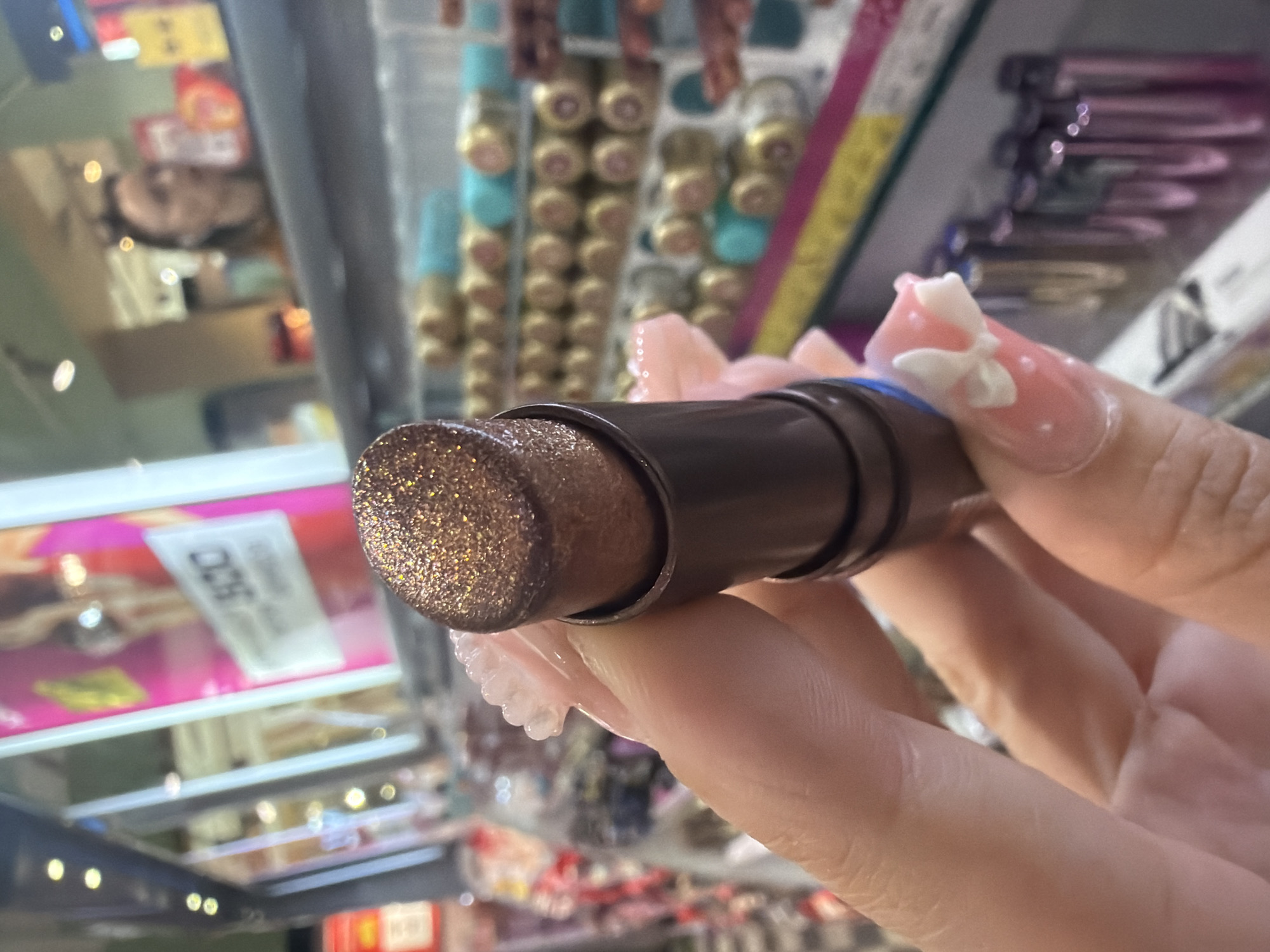An extreme close-up shot focuses on a hand holding a sparkly brown lipstick. The lipstick is angled, revealing its textured, glittery surface. The hand has manicured nails with pink polish adorned with white bows and small white dots. In the background, shelves are stocked with various cosmetic products, including rows of colorful lipsticks or glosses in shades of teal, gold, and purple. The background is blurred, suggesting a retail environment like a makeup store. The lighting appears to be artificial, possibly from overhead store lights, creating a slight glare on some products. The overall scene suggests a personal interaction with a makeup product, likely in a store setting. No specific location in Saint Petersburg, Russia is identifiable from the image.