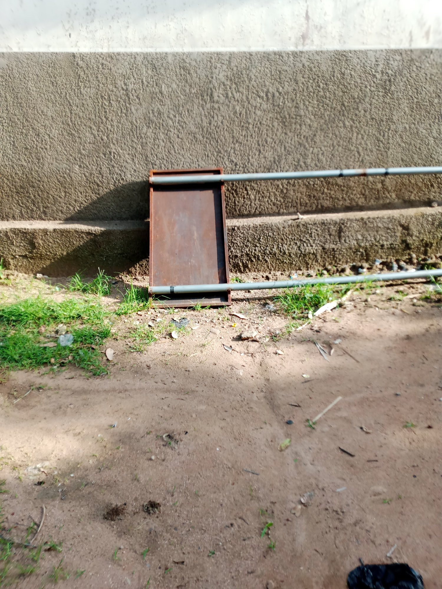 The image depicts an outdoor scene with a textured, greyish-white wall in the background, possibly a building facade. Below the wall is a rough, concrete ledge or step, also textured and grey. In front of the ledge, a brown, rectangular metal object, possibly a shallow box or tray, is placed vertically, supported by two grey pipes that run horizontally across the scene. The ground in the foreground is a mixture of dirt and patchy green grass, with some scattered debris including leaves and small pieces of plastic. The lighting suggests it is daytime, with shadows cast by the wall and the metal object. The environment appears to be an outdoor area in a city, specifically noted as Jauro Yinu, Nigeria.