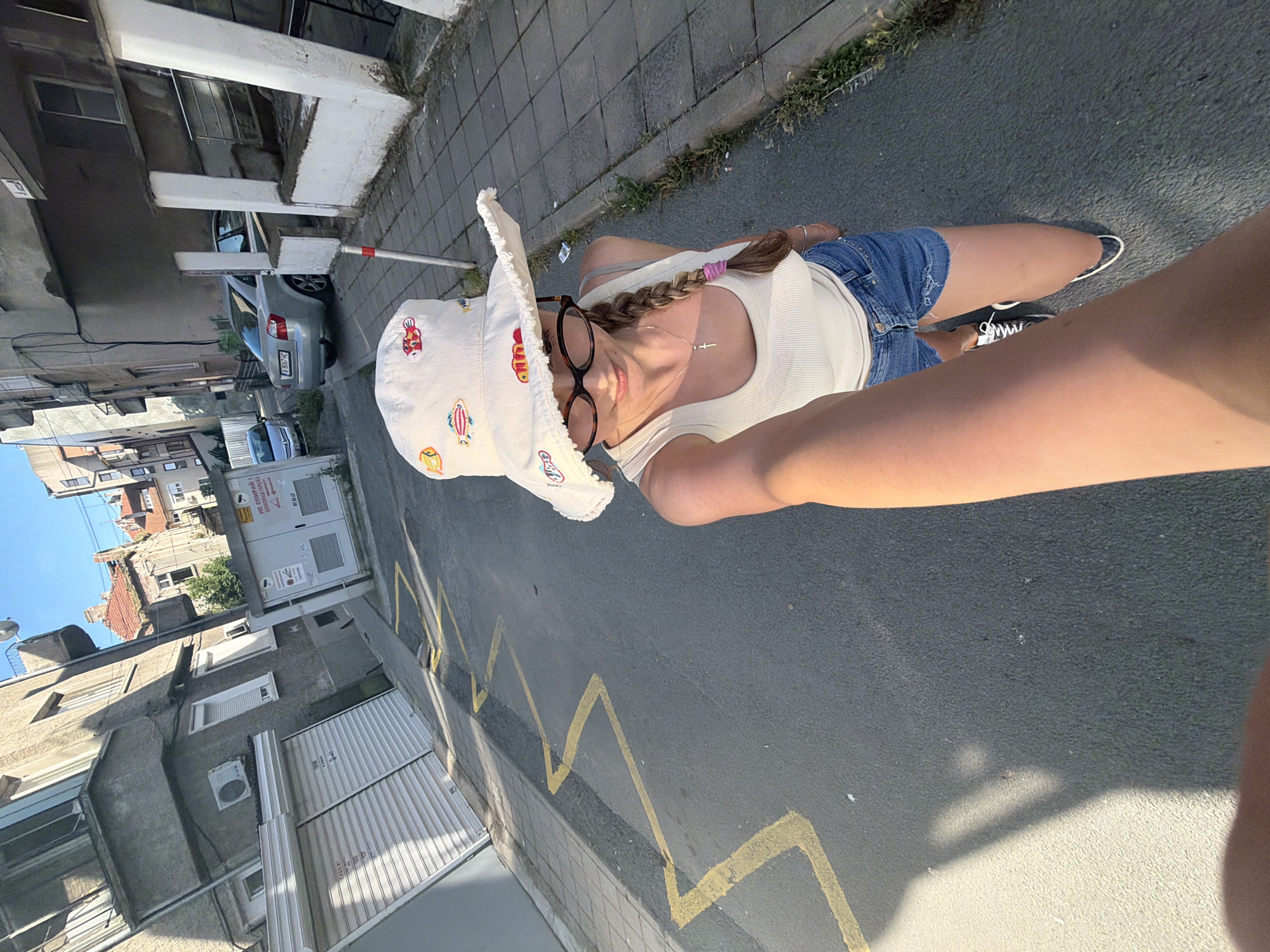 In the city of Burgas, Bulgaria, a young woman with a braided hairstyle and a white bucket hat adorned with embroidered fish, takes a selfie on a paved street. She wears a white ribbed tank top and denim shorts. Her left arm is extended, holding the camera. The street has yellow zigzag markings painted on it, indicating a parking restriction. In the background, buildings of varying architectural styles line the street, some with balconies and shutters. A silver sedan is parked on the right side of the street, and a metallic gray car is visible further down on the left. A utility box with signage is also present. The scene is captured during the day with sunlight casting shadows on the pavement.