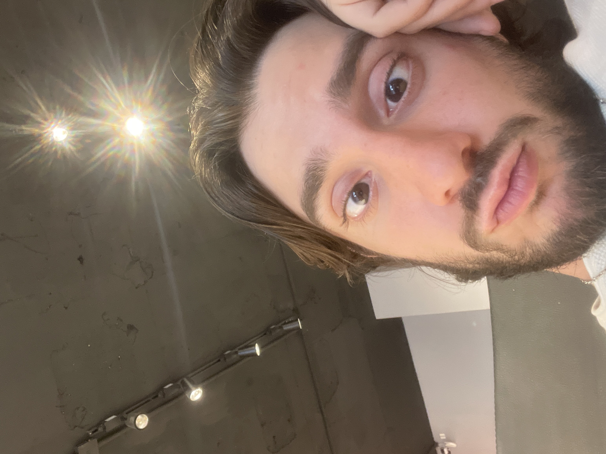A close-up, tilted selfie of a young man with dark hair and a full beard. He is indoors, likely in Mexico City, Mexico, given the lighting fixtures in the background. The wall behind him is gray and has a track lighting system with several small spotlights. The selfie captures the man's face from above, focusing on his eyes, nose, and mouth. His right hand is slightly visible near his forehead. The lighting in the room is a mixture of natural light and artificial light from the spotlights, creating a bright flare in the upper left corner of the image.