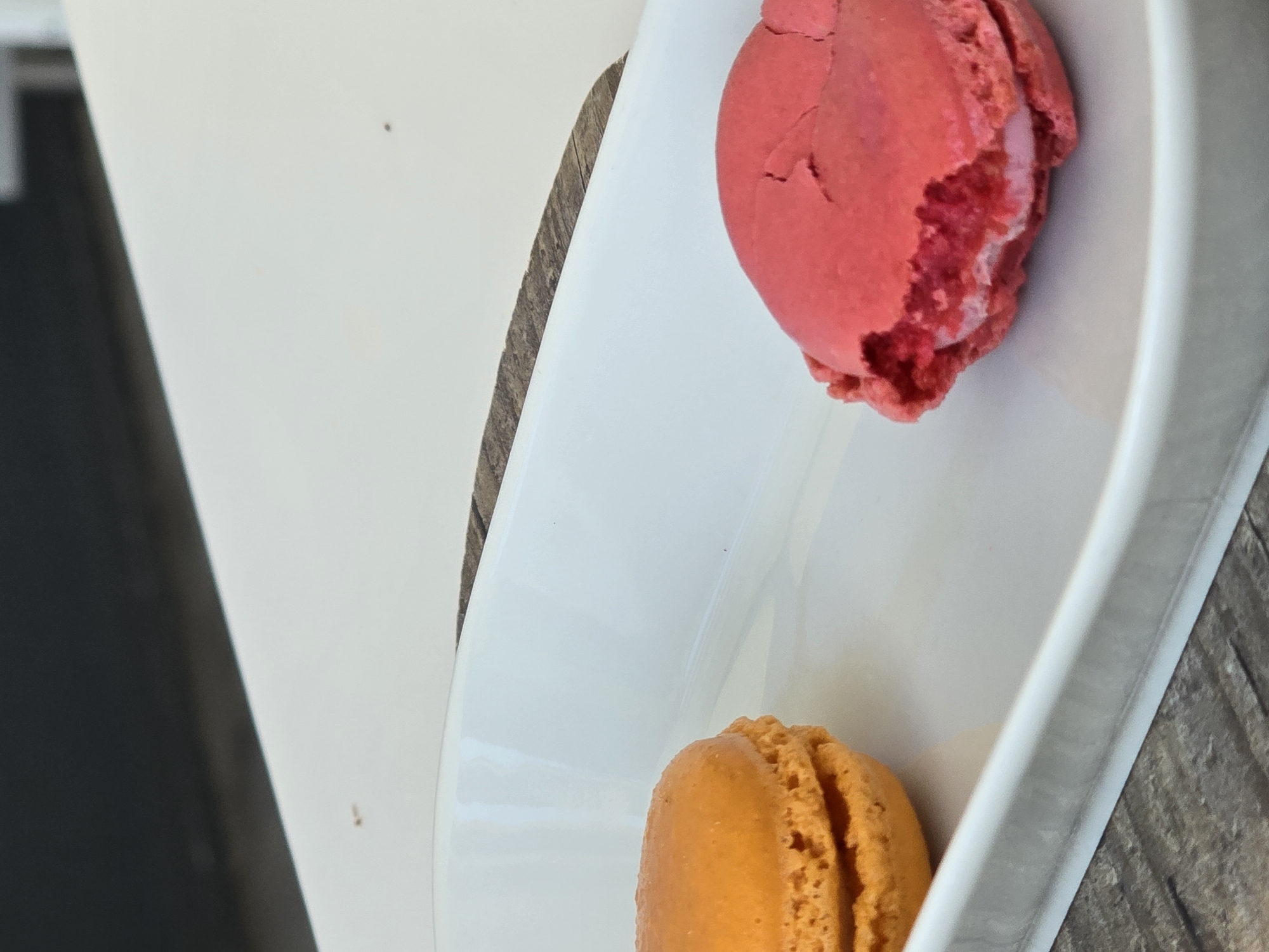 Two macarons are on a white serving tray. The macaron on the left is orange and whole. The macaron on the right is pink and has been partially eaten. The serving tray is on a wooden surface. The background is out of focus and appears to be a dark surface above a light surface. This image was taken in Baronissi, Italy.