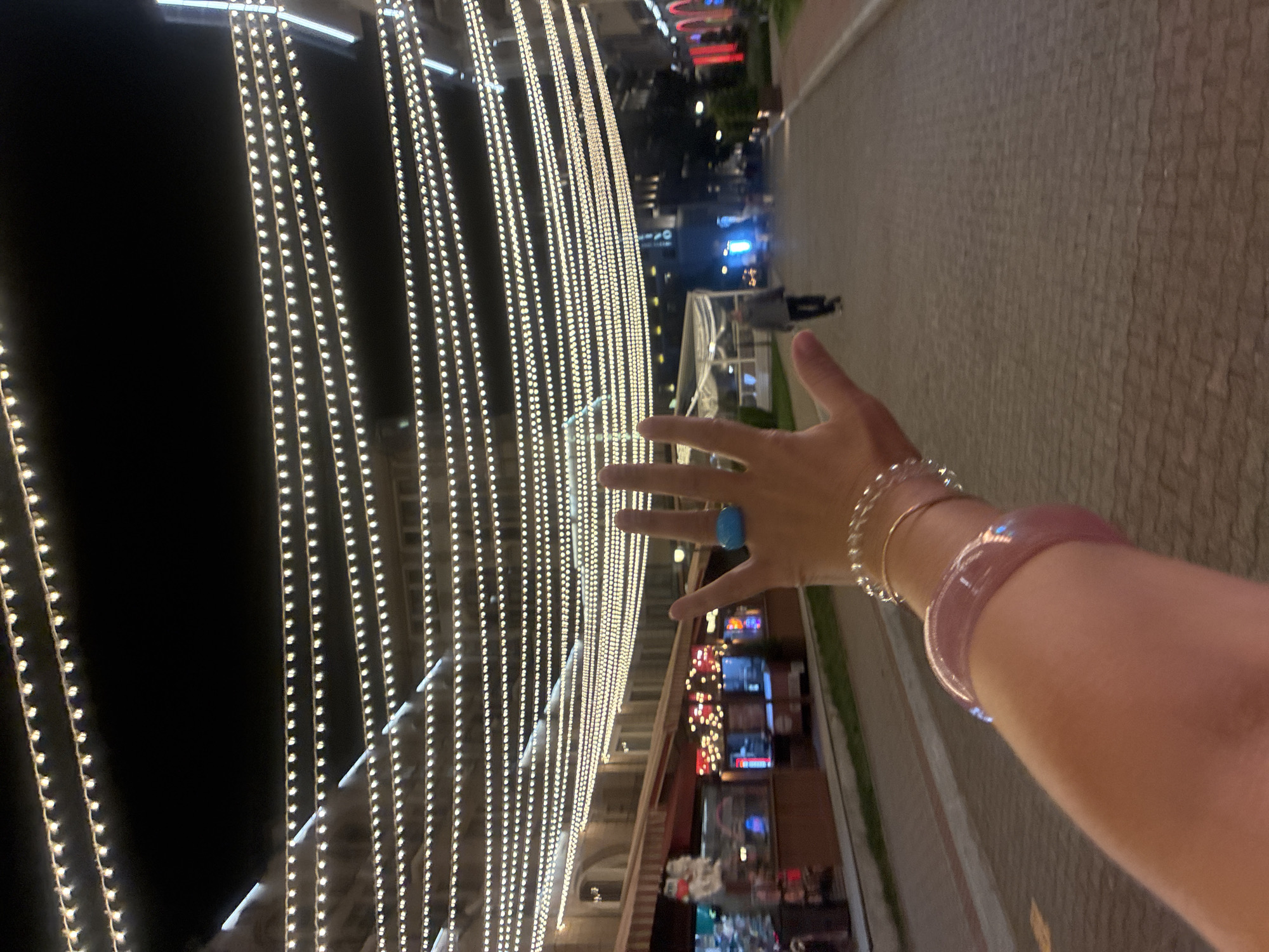 The image depicts a street scene at night, likely in Estosadok, Russia, illuminated by numerous strands of string lights suspended overhead. In the foreground, a person's arm is visible, adorned with several bracelets. One bracelet, a wide, translucent pink band, is positioned closer to the wrist. Above this, a delicate, thin bracelet with clear beads is also visible. On the hand, which is raised and partially obscuring the view of the background, a turquoise ring is worn on the index finger. The hand displays an open palm, with fingers extended, forming a peace sign with the middle and ring fingers.
The background reveals a paved walkway or street lined with buildings. Some of these buildings appear to be shops or restaurants, with awnings and lit displays visible. Further down the street, a person is walking away from the camera, silhouetted against the ambient light. The overall atmosphere is festive and urban, enhanced by the decorative lighting.