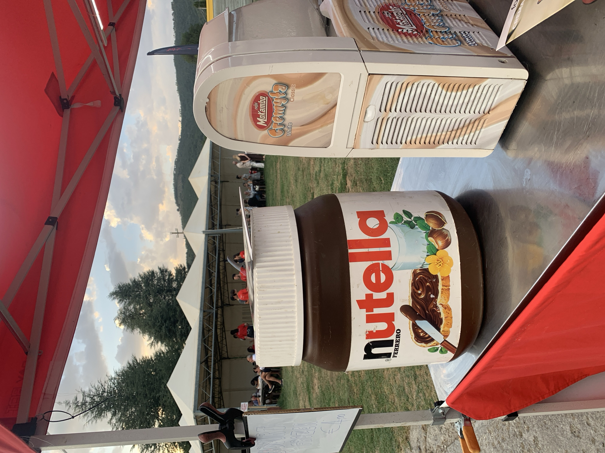 An outdoor scene in Cortona, Italy, features a stall with a large jar of Nutella and a commercial ice cream machine. The stall is covered by a red canopy. In the foreground, a large jar of Nutella is prominently displayed next to a cream-colored ice cream machine with branding for "Mokambo Cremy'ta". A red cloth drapes over the edge of the stainless steel table where these items are placed. To the left of the Nutella jar, a white sign attached to a pole has handwritten text including "FROZEN" and "€4.00". In the background, several white tents are set up, with people gathered around tables and walking around. The sky above is partly cloudy, and a wooded hillside is visible in the distance.