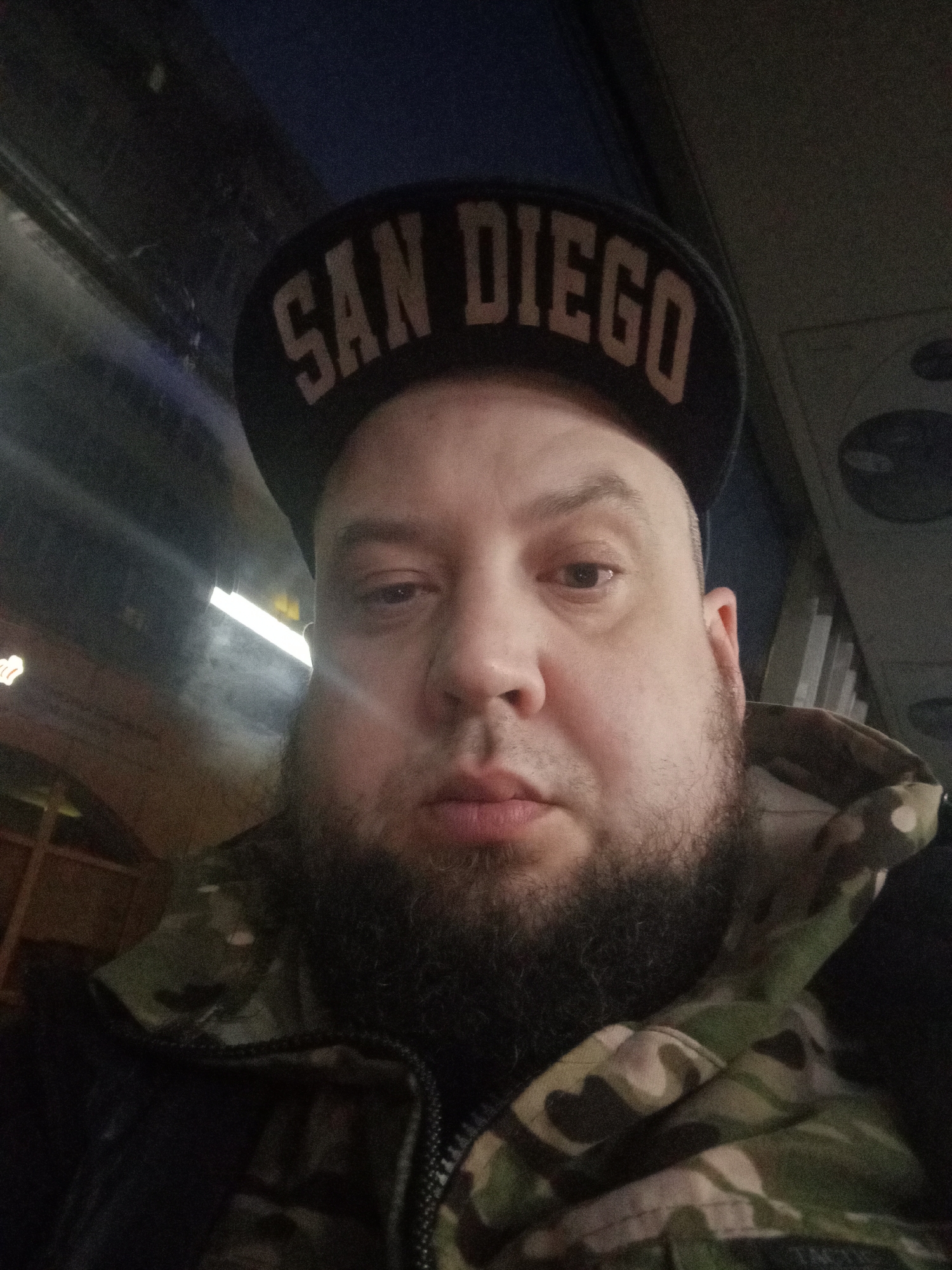 A medium close-up photograph depicts a man with a full beard wearing a "SAN DIEGO" baseball cap and a camouflage jacket. The lighting is dim, suggesting an indoor setting. The background appears to be a public transportation interior, possibly a bus or train, given the overhead structures and window reflections. The image does not contain sufficient detail to determine whether it was taken in Neryungri, Russia.