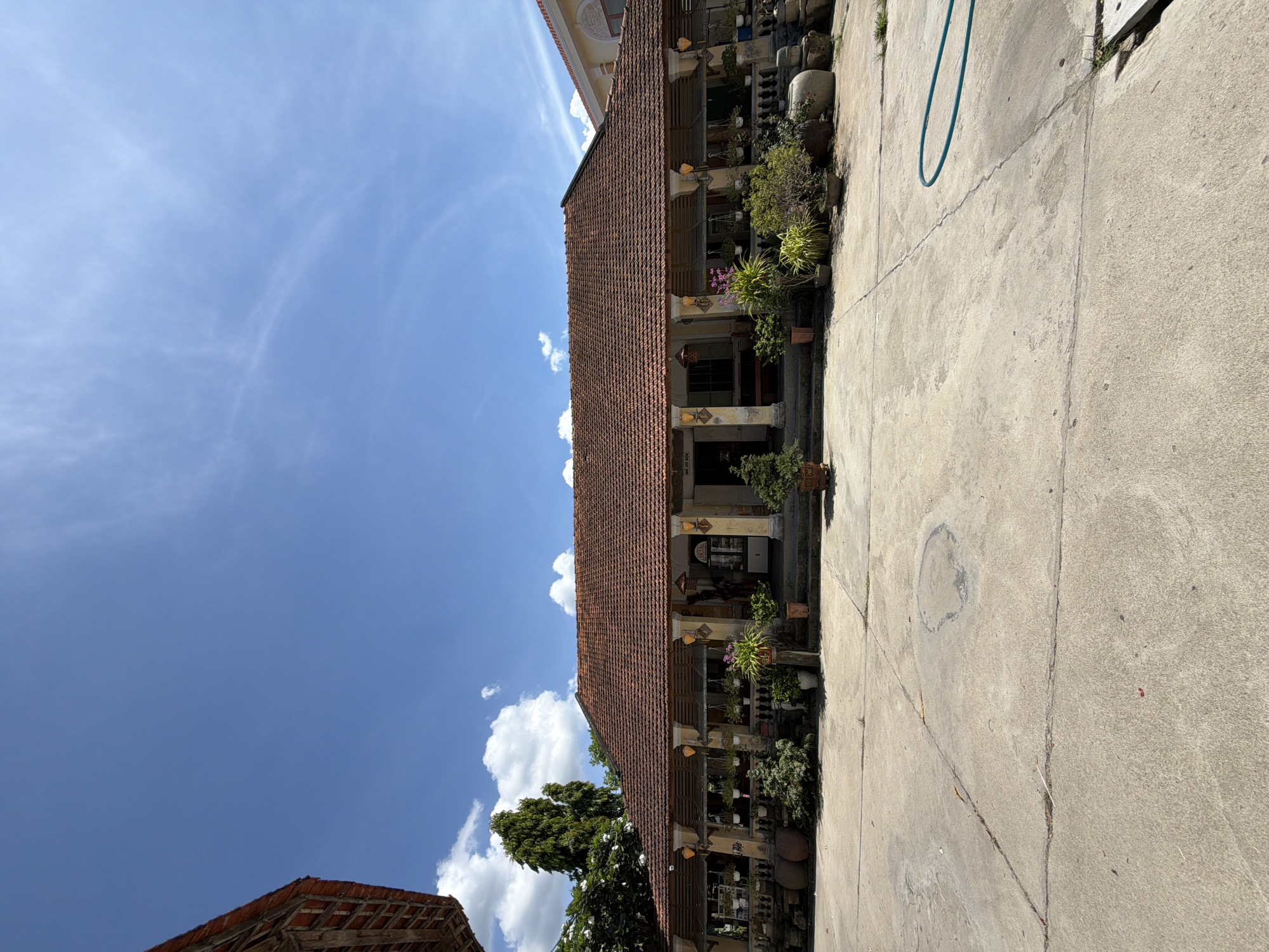 This is a wide-angle, eye-level shot of a traditional Vietnamese house on a sunny day. The house, likely located in Hội Tín, Vietnam, features a red-tiled, pitched roof with exposed wooden beams on the eaves. The facade is light yellow, with a covered porch supported by four fluted columns. The porch is lined with decorative pottery, planters filled with various greenery and flowers, and hanging baskets. A set of stairs leads up to the entrance, which is partially obscured. A large tree with lush foliage is visible to the left of the house, and another building with a similar roofline is seen to the right. The foreground is a concrete courtyard with visible cracks and stains, and a teal-colored garden hose lies coiled on the ground. The sky is bright blue with scattered white clouds. There are no people or animals visible in the frame.