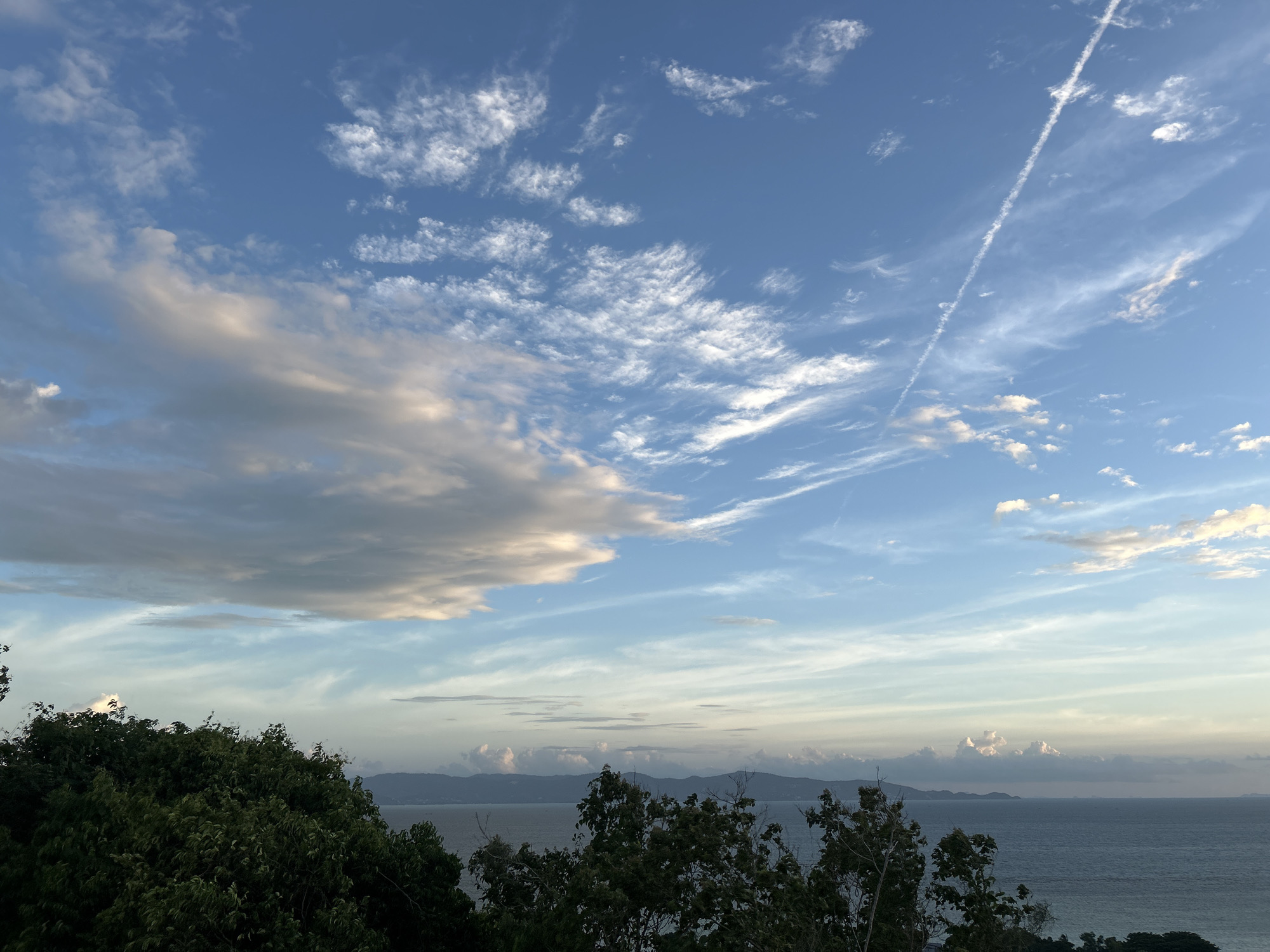 The image depicts a serene coastal landscape in Ko Pha-ngan District, Thailand, likely taken from an elevated viewpoint. The scene is dominated by a vast expanse of sky and sea. The sky is a gradient of blues, transitioning from a deeper hue at the top to lighter shades near the horizon. Wispy clouds are scattered across the sky, with some appearing feathery and others more substantial and tinged with the warm colors of either sunrise or sunset. A distinct contrail from an aircraft is visible diagonally across the upper right portion of the sky.
Below the sky, the sea stretches out, appearing calm with subtle ripples. In the distance, a line of silhouetted mountains forms the horizon, separating the sea from the sky. The foreground is characterized by a dense canopy of lush green trees, their foliage dark and rich, partially obscuring the view of the sea and mountains. The overall impression is one of tranquility and natural beauty, characteristic of a tropical island setting. There are no people or specific man-made structures clearly visible in the image, emphasizing the natural environment.