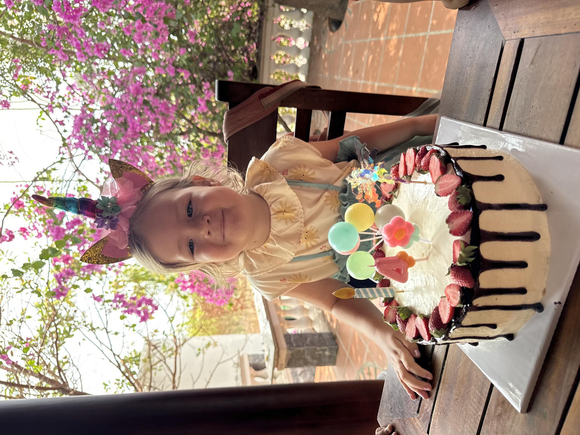 This media file captures a festive outdoor birthday celebration, likely in Mũi Né, Vietnam. The central figure is a young girl, possibly around four years old, indicated by the prominent number '4' candle on her cake. She has fair skin, blonde hair, and is wearing a light-colored top adorned with sun motifs, along with what appears to be a light blue skirt or dress. A colorful unicorn headband, featuring a rainbow horn, glittery gold ears, and pink and purple floral details, adorns her head. She looks directly at the camera with a gentle, content smile, radiating happiness.
In front of the girl, resting on a rustic wooden table, is a round birthday cake. The cake is covered in smooth white or cream frosting, elaborately decorated with fresh, halved strawberries lining its circumference and rich chocolate drizzles cascading down its sides. Alongside the "4" candle, the cake is further embellished with whimsical toppers including small, colorful balloons in shades of yellow, green, and pink, a light pink flower, and an orange heart shape.
The background features a vibrant display of pink bougainvillea flowers, suggesting a lush, warm, and tropical environment. Hints of a tiled architectural structure are also visible, likely part of a patio or outdoor living space. The scene is brightly lit by natural daylight, contributing to the cheerful and celebratory ambiance. The overall impression is one of joy and youthful celebration.