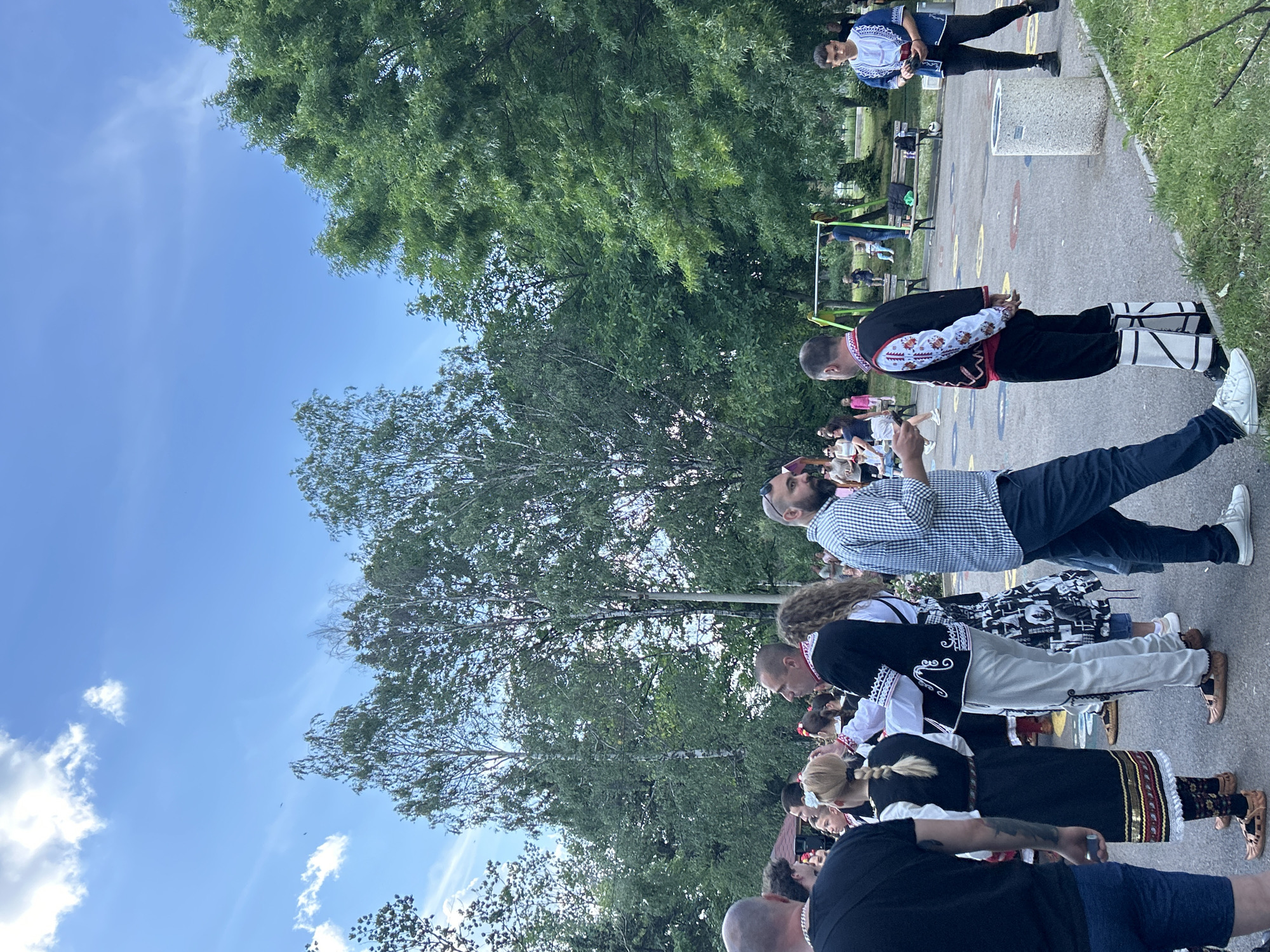 A group of people are standing together in Mirovyane, Bulgaria, near trees and a blue sky with clouds. Some people are wearing traditional Bulgarian clothing.