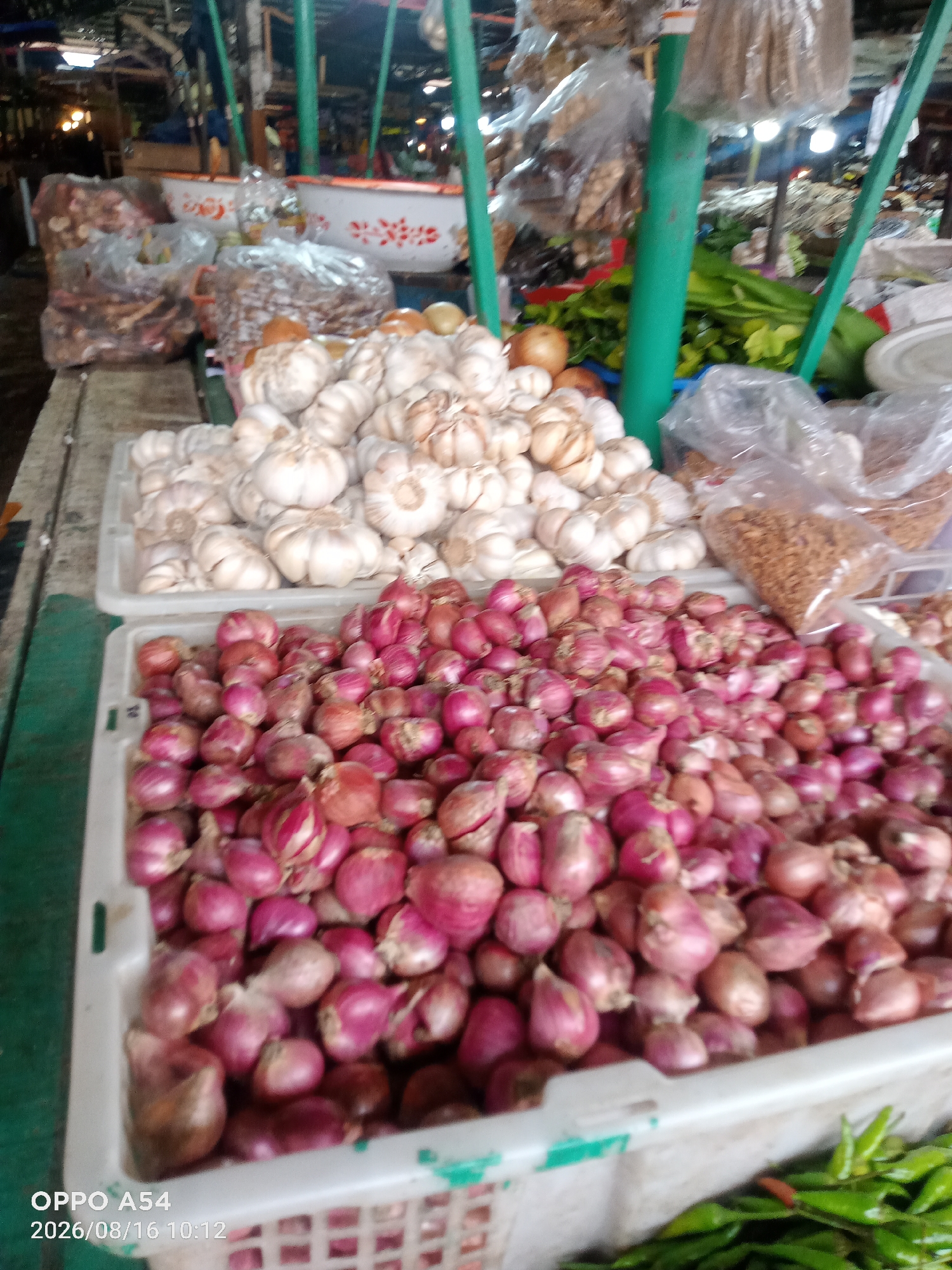An outdoor market stall in Jambi, Indonesia, displays a variety of produce. In the foreground, a white plastic basket is filled with shallots. Behind the shallots, another white plastic basket holds garlic bulbs. To the right of the garlic, a clear plastic bag contains dried spices or seeds, and further back, a bundle of green leafy vegetables is visible. In the background, the market structure with green support poles and a roof is apparent, with other stalls and goods blurred in the distance. The lighting suggests it is daytime.
