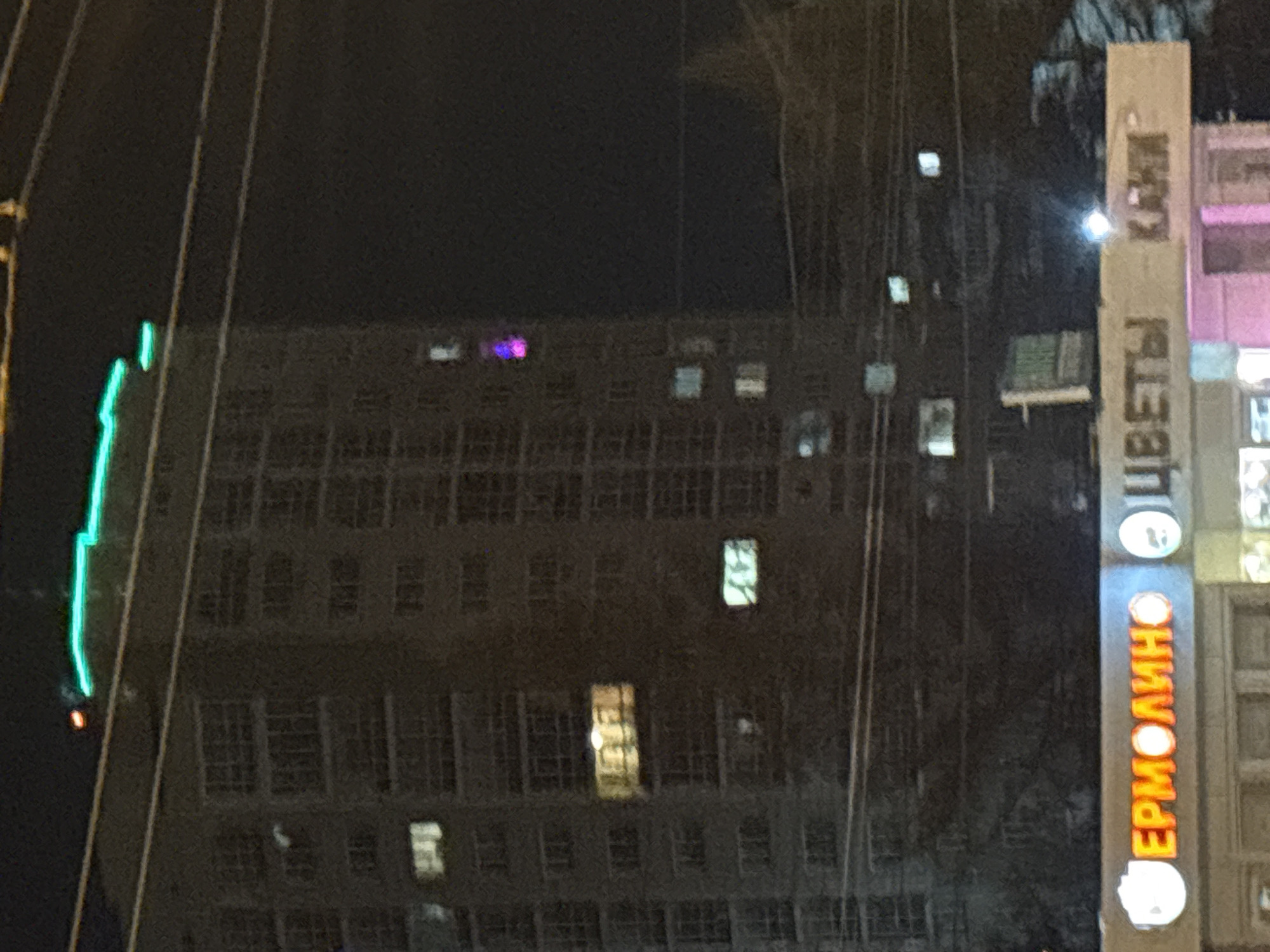 The image depicts a nighttime urban scene in Krasnoyarsk, Russia. An apartment building with illuminated windows occupies the left and center. To the right, vertical signs read "ЕРМОЛИНО" and "ЦВЕТЫ КАКИМ". Electrical lines are visible across the image. There are no people visible.