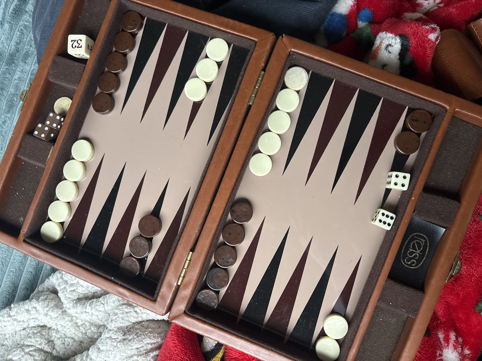 The image depicts an open backgammon board with a brown and beige interior. The left side has five dark brown pieces in the upper right corner, followed by five white pieces lower down, and five dark brown pieces in the bottom right corner. A doubling cube with the number "32" is placed in the upper left corner of this side. Additionally, two dice are placed in the lower left corner of this side. The right side features five white pieces in the upper left corner and two dark brown pieces in the upper right corner. At the bottom of the right side, there are three white pieces. Two dice are also visible near the lower right corner of this side. The backgammon board is surrounded by fabric, including a red and white patterned material. There is no evidence to identify that this image was taken in Puebla, Mexico.