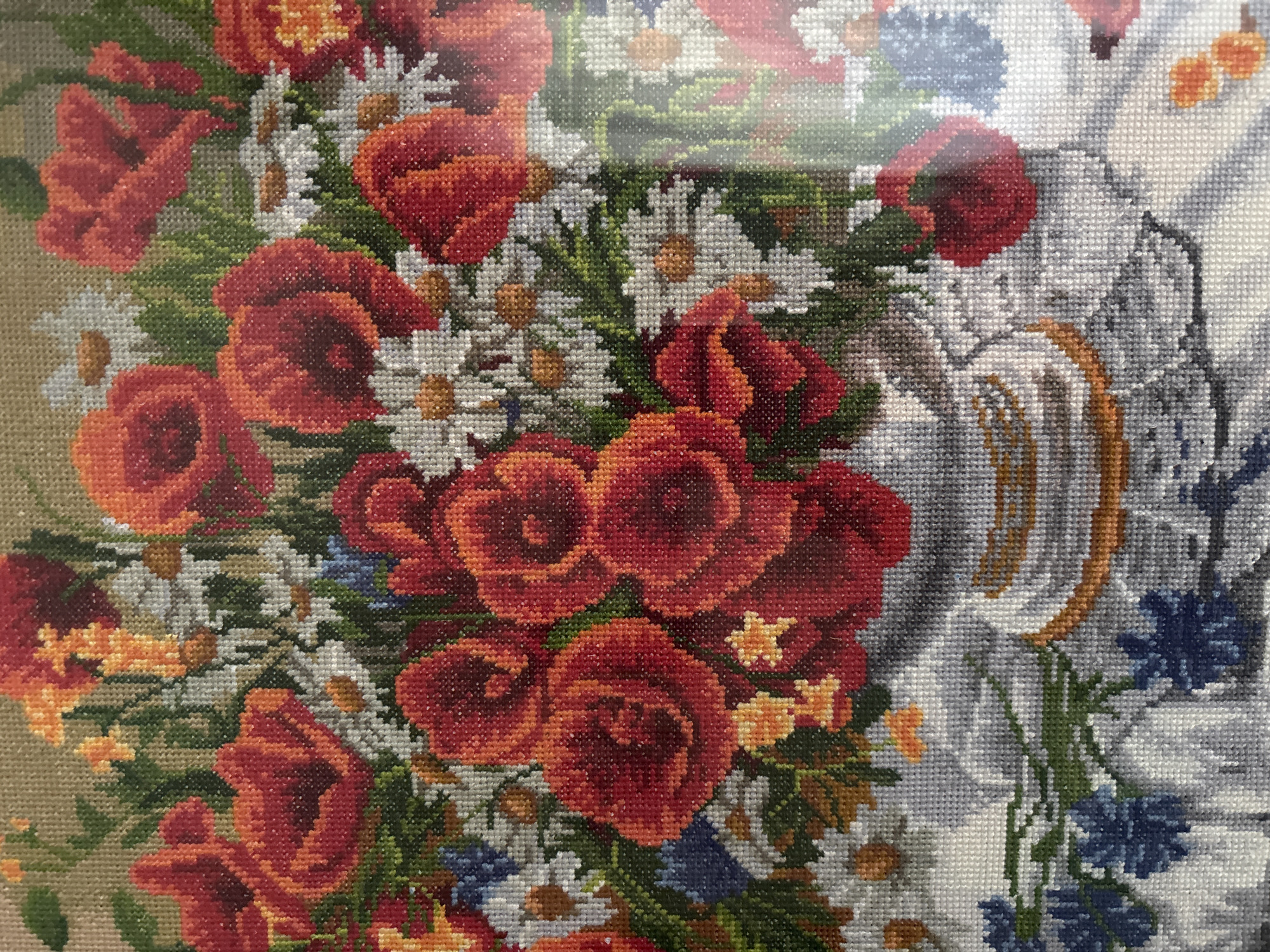 This is a close-up, cropped image of a cross-stitch embroidery depicting a vibrant bouquet of red poppies and white daisies in a silver vase. The arrangement also includes hints of orange and blue flowers and green foliage. The vase sits on a surface covered with a white, textured cloth, possibly a tablecloth. The background is a neutral, woven texture. The image is a detail of a larger embroidery, and due to the cropped nature, specific details like the location within the city of Tomsk, Russia, cannot be determined. There are no people or actions depicted.