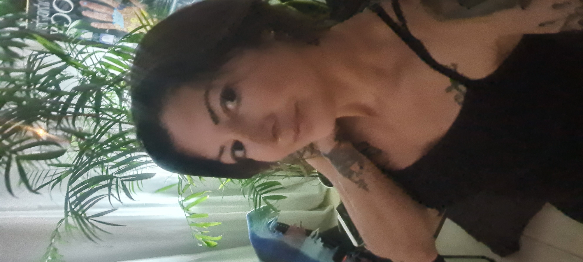 A person is visible, positioned against a backdrop of green foliage, possibly an indoor plant. The person has dark hair, light skin, and is wearing a black top. There is visible tattooed skin on the arm. The context of Nahariyya, Israel, is not discernible from the image.