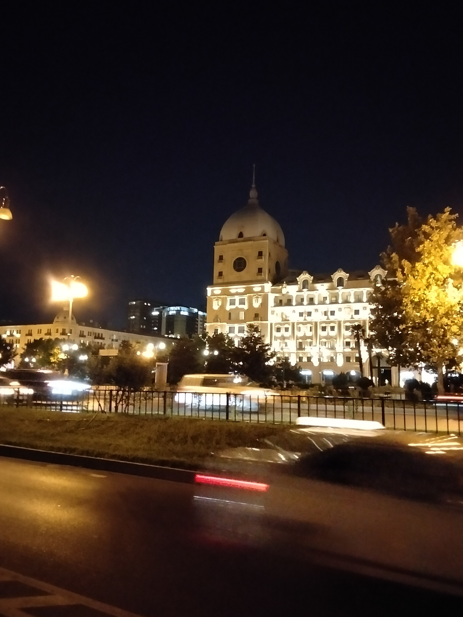 The image depicts a nighttime scene on a street in Baku, Azerbaijan, with a prominent building featuring a large dome and illuminated windows. The architecture of the building suggests a historical or significant structure, possibly a government building or hotel. In the foreground, a street is visible with blurred streaks of light indicating moving vehicles, suggesting a long exposure photograph. A fence separates the street from a grassy area and trees. The sky is dark, indicating it is nighttime. Another building, also illuminated, is visible to the left, as well as streetlights. The overall impression is of a city at night, with a blend of historical and modern elements, and a sense of urban activity.