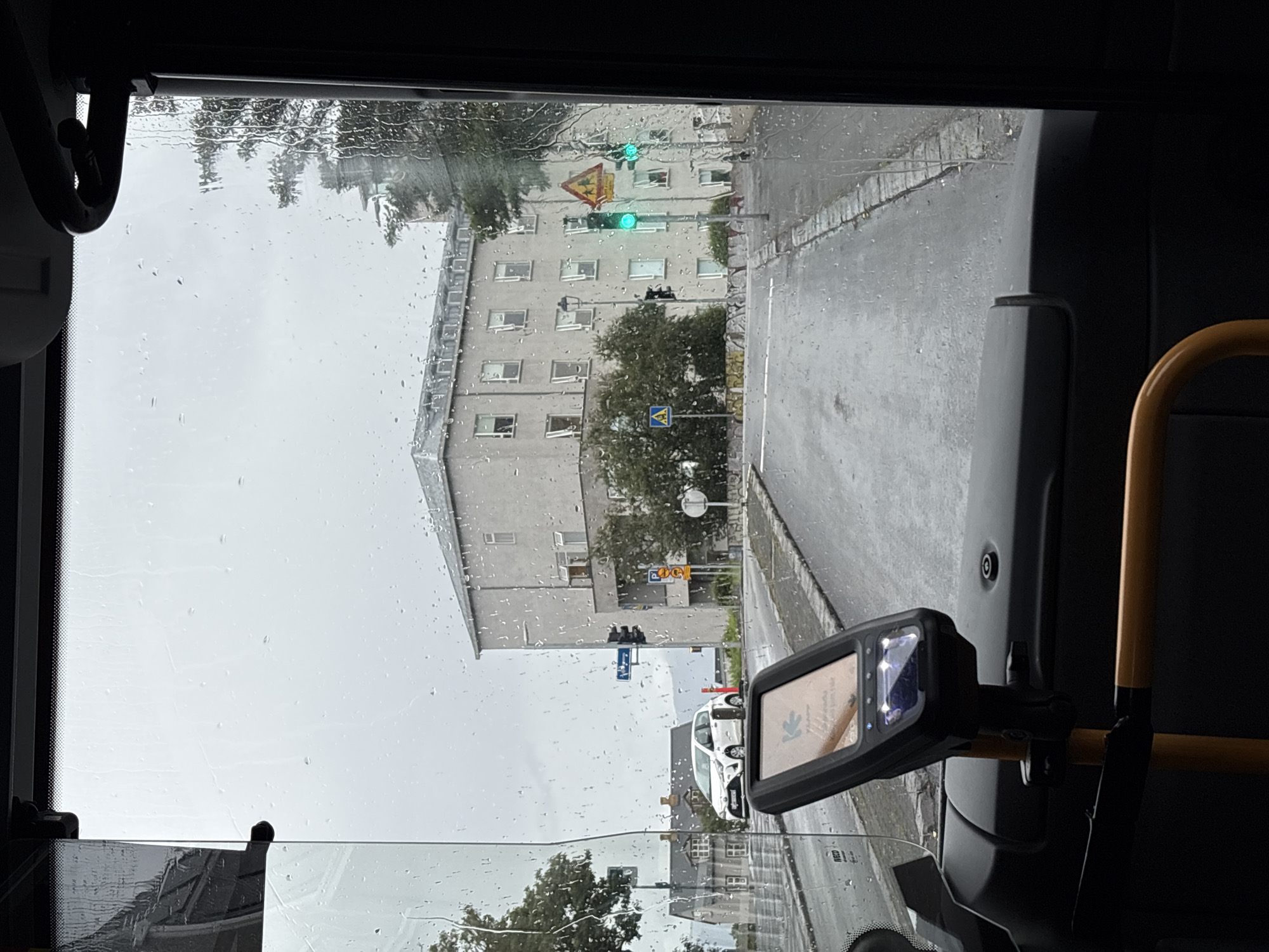The image shows a view from inside a bus in Reykjavik, Iceland, during a rainy day. The windshield is covered with raindrops, distorting the view of the outside. In the foreground, a ticket machine is mounted on the dashboard. Outside, a grey, multi-story building with several windows is visible. A white car is parked on the wet road. Traffic lights show a green signal, and various street signs are present, including a blue directional sign and a yellow warning sign with children crossing. The road appears to be a city street in Reykjavik.