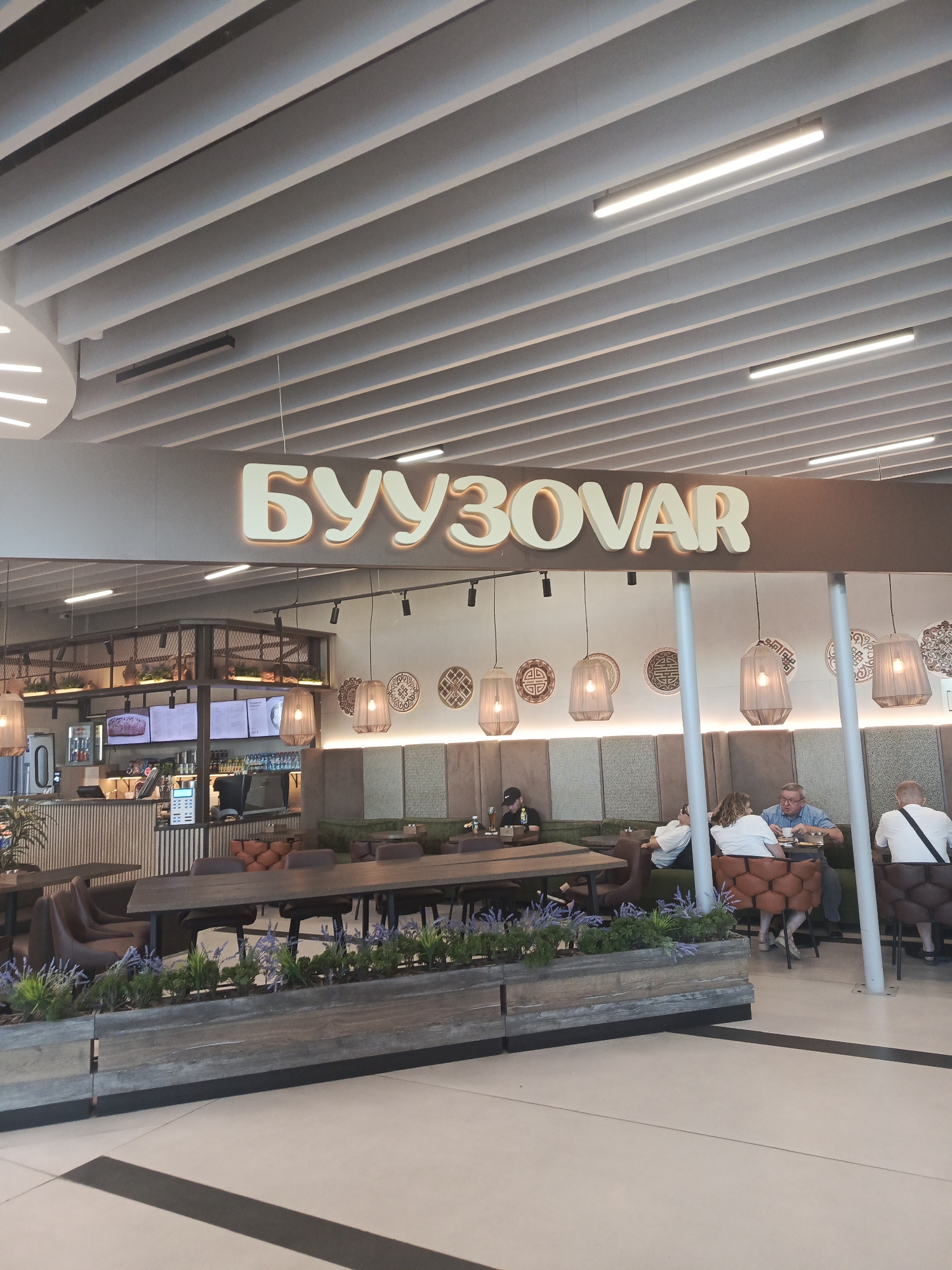 The image shows the interior of a cafe, identified by the signage "БУУЗОВАР" (BUUZOVAR) located in the city of Ob, Russia. The cafe features a modern design with a slatted ceiling and contemporary lighting. Several patrons are seated at tables, engaged in conversation and dining. The seating area includes booths and individual tables, with some patrons dressed casually. The cafe appears to be well-lit and decorated with ornamental plates on the wall and hanging lamps. A counter area with a menu display is visible in the background. The overall atmosphere suggests a public dining establishment.