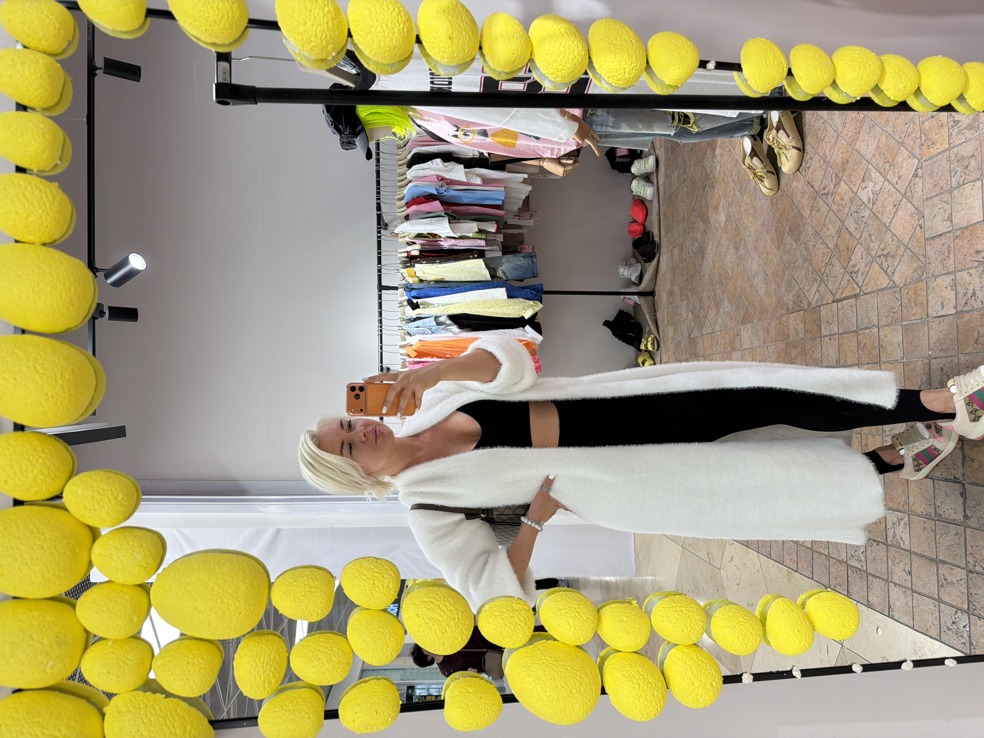 A woman with short blonde hair is posing for a mirror selfie, holding a phone in her right hand. She is wearing a black crop top, black leggings, and a long white cardigan. The mirror she is using is decorated with yellow lemon-like objects around the frame. Behind her, clothes are hanging on a rack, and there are also shoes displayed. The floor is tiled. The location appears to be a retail store.