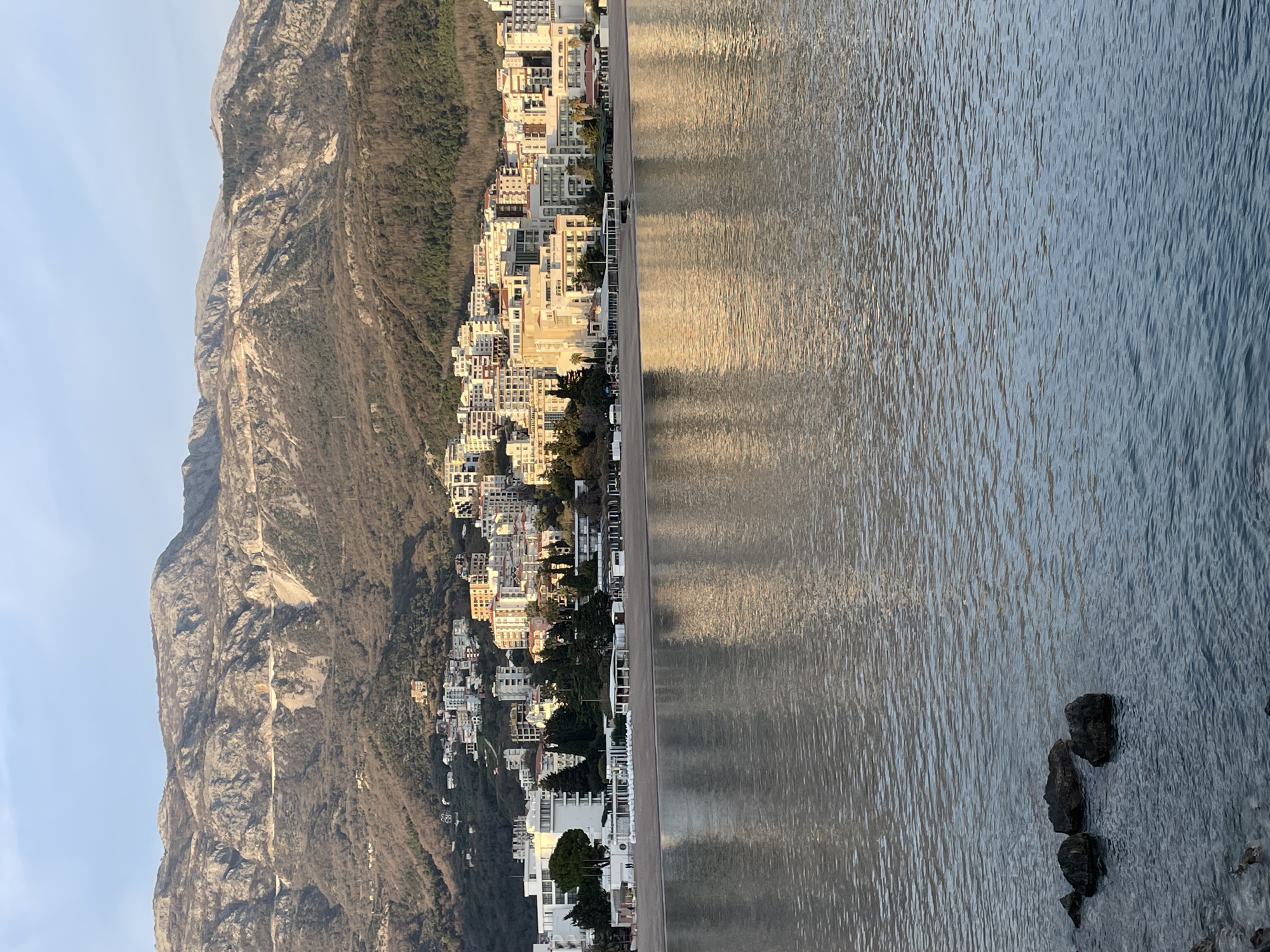 The image presents a panoramic coastal scene of Budva, Montenegro, observed from the water. The time of day appears to be either sunrise or sunset, indicated by the warm, directional light illuminating the cityscape and reflecting on the water.
In the foreground, a body of water exhibits gentle ripples across its surface, reflecting the warm light. Three dark, irregularly shaped rocks are partially submerged in the water in the lower-left area.
The midground features a wide, light-colored sandy beach stretching horizontally. Along the beach's edge, a series of structures are visible: modern, multi-story white buildings with flat roofs, interspersed with establishments featuring covered outdoor areas and awnings. Rows of stacked white objects, consistent with beach furniture such as sun loungers, are present on the sand in several locations.
The background is dominated by a densely developed urban area, characteristic of Budva, which ascends the lower and mid-slopes of a large mountain range. The city consists of numerous multi-story buildings of varying architectural styles and heights, predominantly light-colored, with some exhibiting warmer, possibly terracotta, hues. Trees and sparse vegetation are visible between and around the buildings. Behind the urban development, rugged mountains rise prominently. Their slopes display rocky outcrops, steep inclines, and patches of sparse, dry vegetation. A winding road is discernible traversing a mid-level section of the mountain face. The sky above is clear and light blue. No distinct individuals or specific human actions are discernable within the frame.