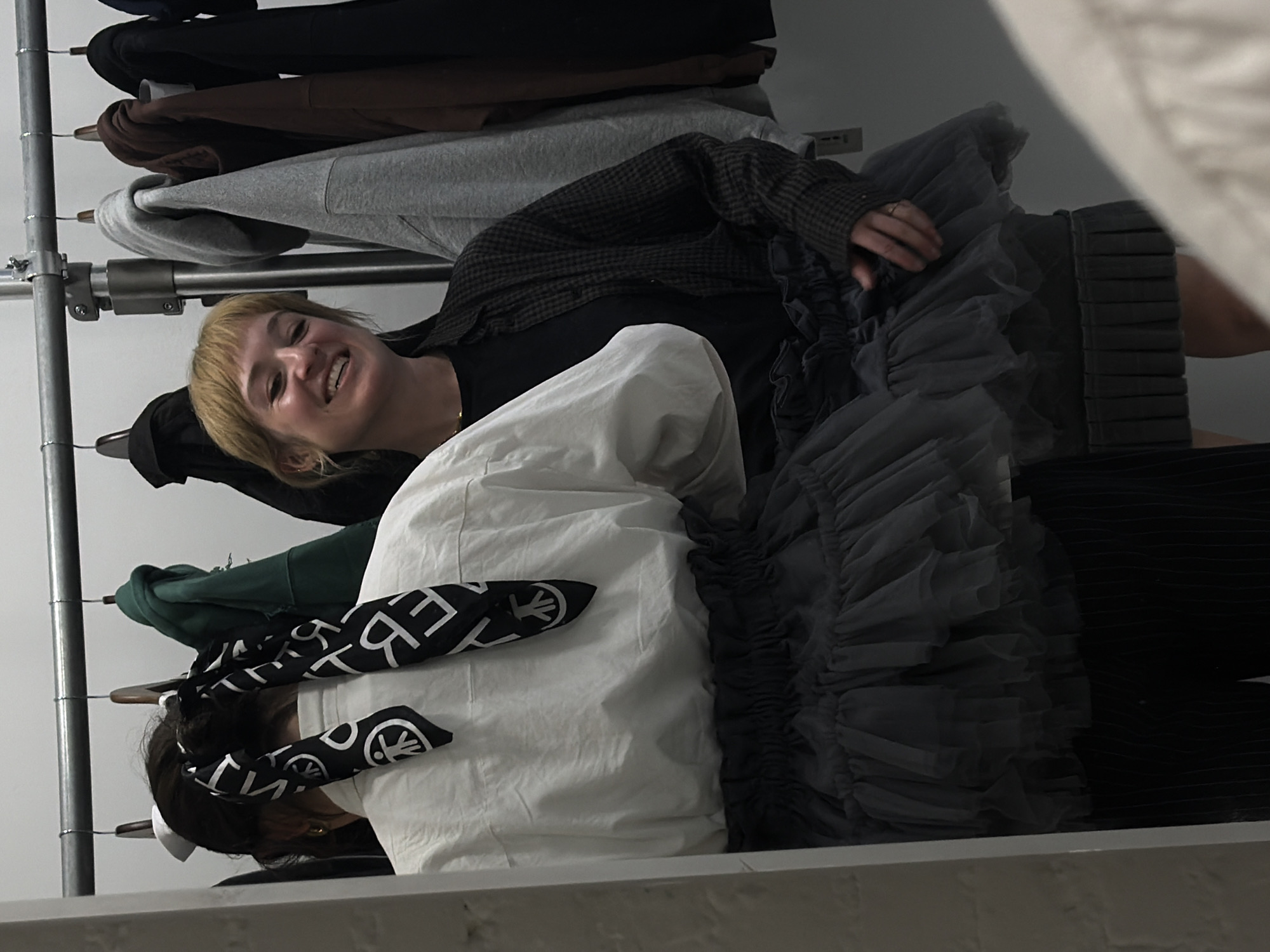 The image shows two women in what appears to be a clothing store or backstage area. One woman, with short blonde hair, is smiling and looking towards the camera, wearing a dark shirt and a patterned blazer. The second woman, with dark hair tied back with a patterned black and white scarf, is partially obscured behind a white garment. A clothing rack with various garments including hoodies and jackets is visible in the background. The environment appears to be an indoor retail or fashion setting, but the specific location within New York City cannot be determined from the image.