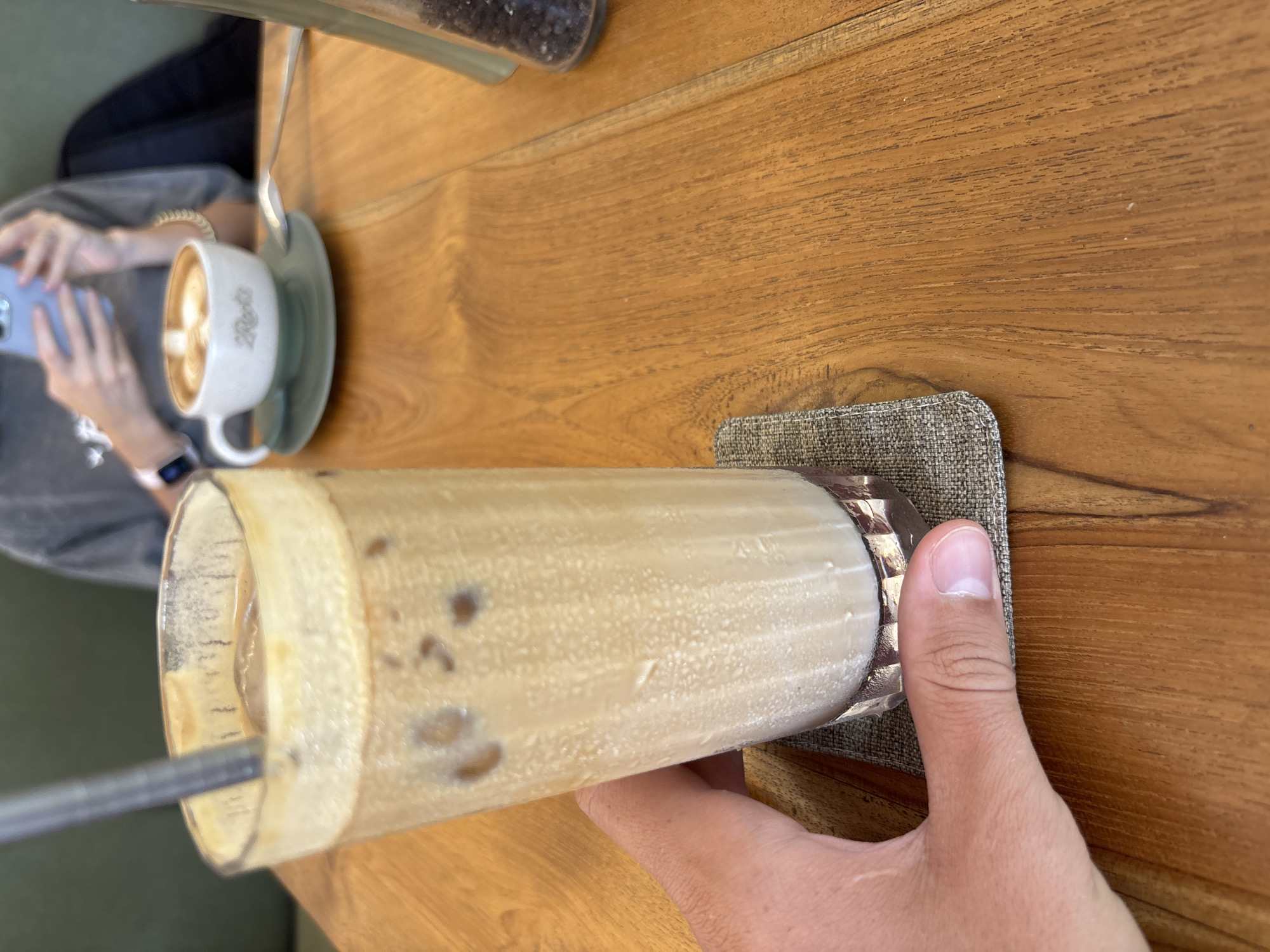 A high-angle, eye-level, indoor shot shows a tan wooden table with an iced latte in a tall glass being held by a hand.
The hand grips the lower portion of the glass. The drink sits on a small, gray woven square coaster. There's a silver straw in the latte. A coffee cup and saucer with a spoon are to the left of the iced latte. A person partially appears, holding a smartphone. A glass container of coffee beans is in the upper left corner.
The location is likely a cafe in Kuta Selatan, Indonesia.