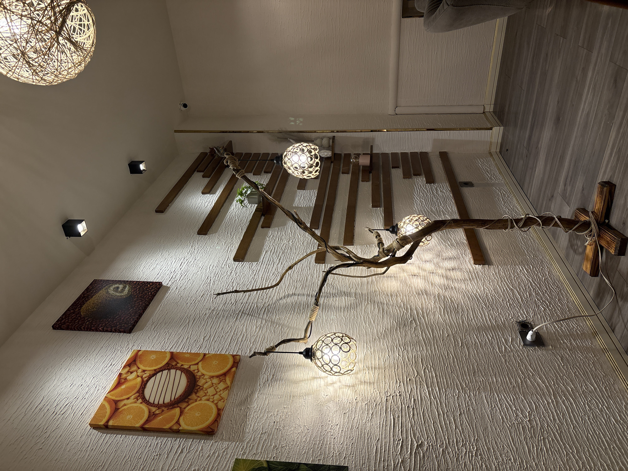 The image depicts an interior scene, likely a cafe or a residential space, with a unique decorative tree-like floor lamp as the focal point. The lamp, made of natural branches, has three globe-shaped lampshades with intricate metalwork. It is placed on a wooden base on a grey laminate floor. The wall behind the lamp is textured white and features two framed pictures: one with sliced oranges and another with a brown swirl pattern. Several staggered wooden shelves are mounted on the wall, some holding small decorative items, including a plant. Two square black ceiling lights are visible, along with a large, spherical, woven pendant light fixture. A security camera is also mounted on the wall near the ceiling. The overall ambiance is warm and inviting, with soft lighting creating shadows and highlighting the textures of the room. There is no indication of the location being in Khatay Raion, Azerbaijan, nor are there any people present in the image.