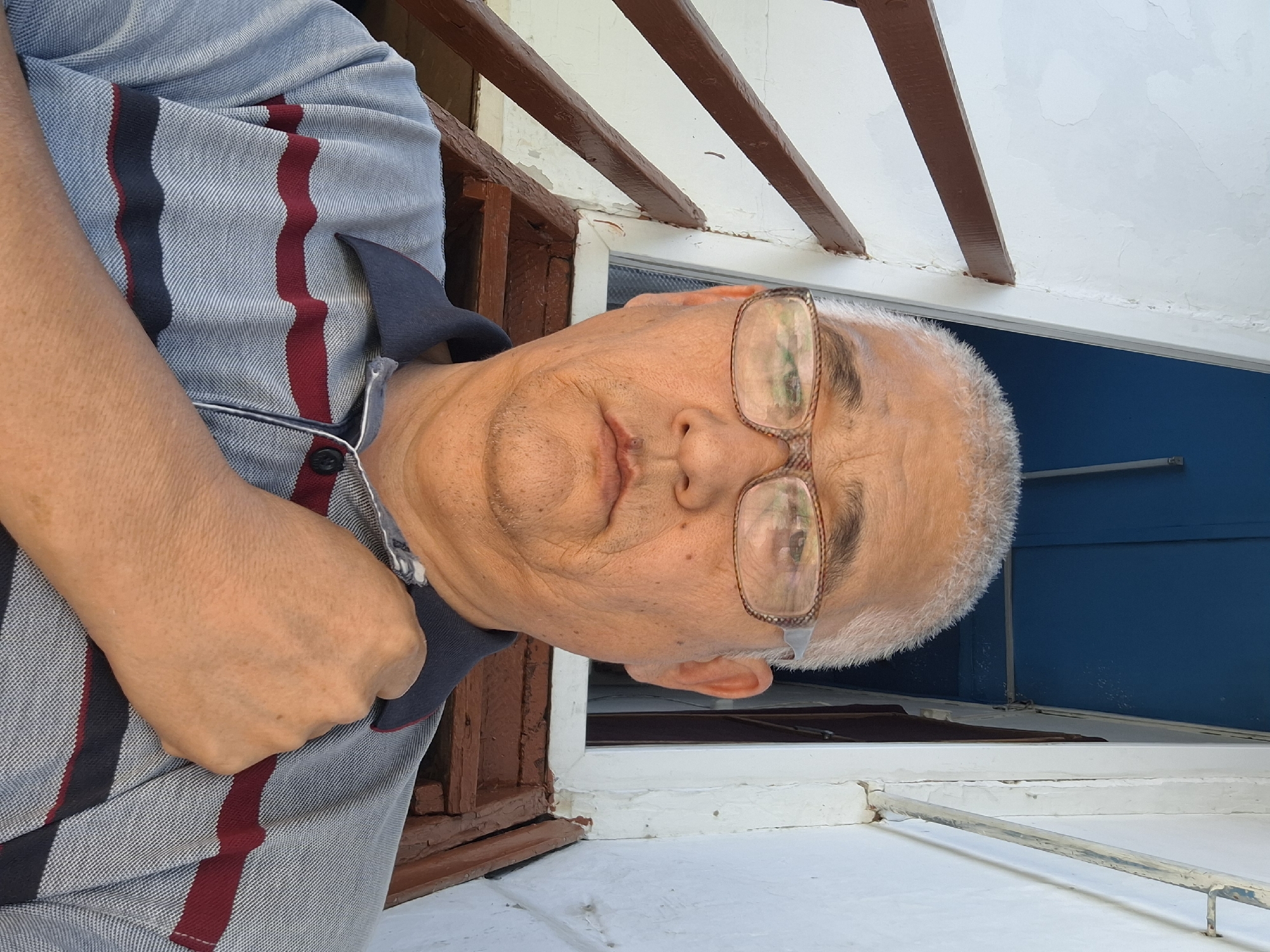 A man with graying hair and glasses is captured in a close-up selfie outdoors. He is wearing a striped polo shirt and has his right hand placed over his chest. Behind him, a wooden staircase with dark brown railings is visible, leading up to a doorway with blue trim. The background suggests an outdoor setting, possibly a residential area in Almaty, Kazakhstan, given the architectural style and natural light. The man's expression is neutral, and he appears to be looking directly at the camera. The overall scene is simple and candid.