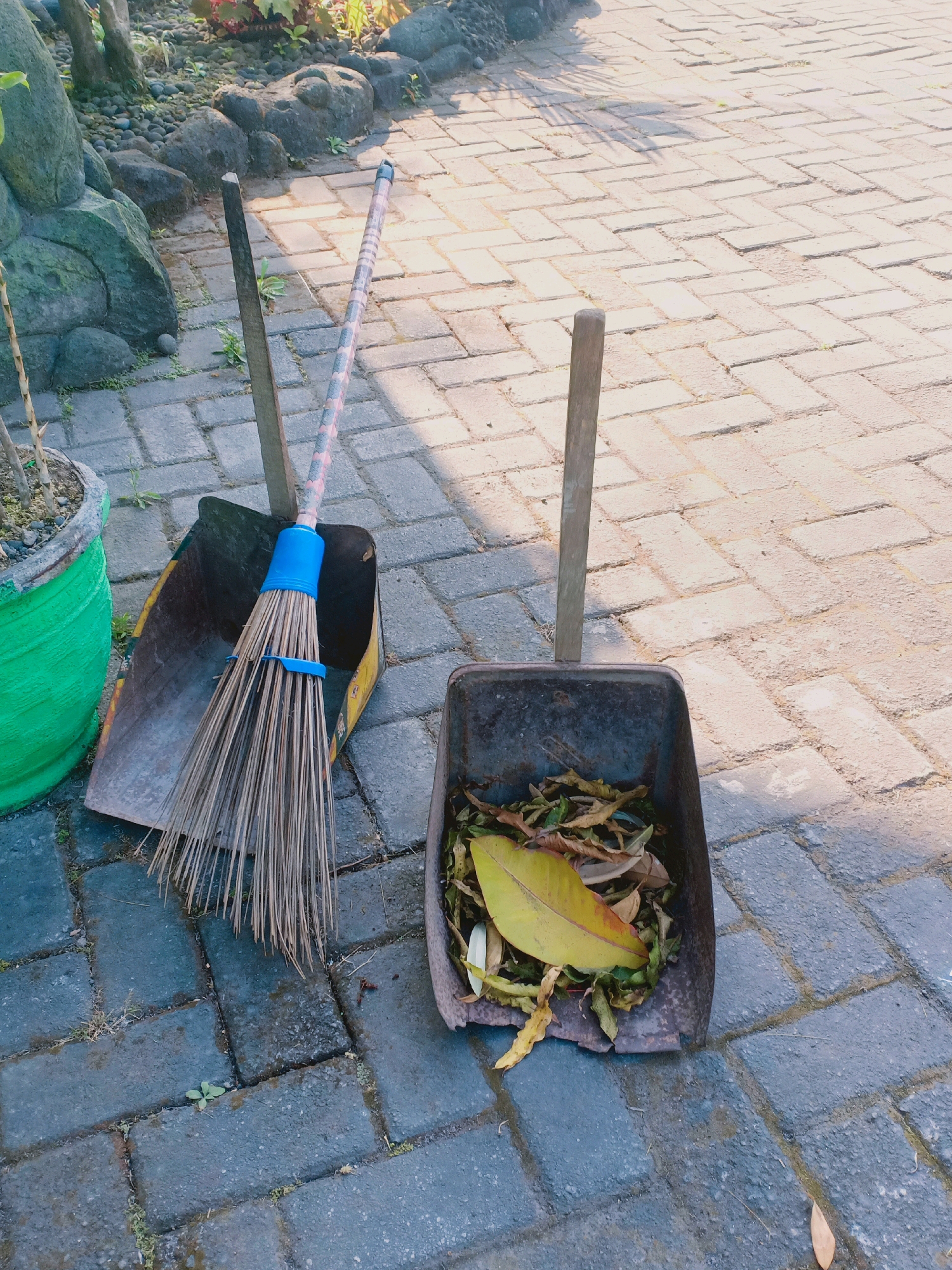 Two dustpans and brooms are placed on a brick walkway. The dustpan on the left contains a broom with a blue band around its bristles, and the dustpan on the right contains fallen leaves. The environment appears to be outdoors, possibly in a garden or courtyard, with a stone structure and a potted plant visible in the background. The brick walkway suggests an urban or suburban setting. The scene is lit by sunlight, casting shadows on the ground. There are no people or animals visible in the image. The overall impression is one of a cleaning in progress or recently completed.