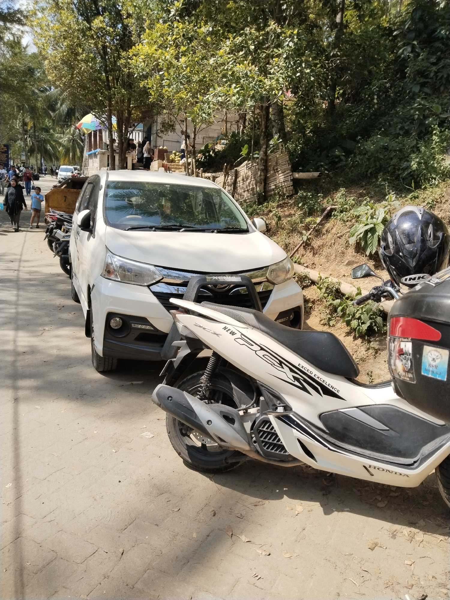 In Prigi, Indonesia, a white Toyota Avanza is parked on a dirt road, facing left. A white Honda PCX scooter is parked to the right of the car, with its front end angled towards the car. A black helmet rests on the scooter's handlebars. In the background, a lush green hillside is visible, with trees and vegetation. Several people are walking along the road, some of whom appear to be tourists. The scene is illuminated by bright sunlight, casting shadows on the road.