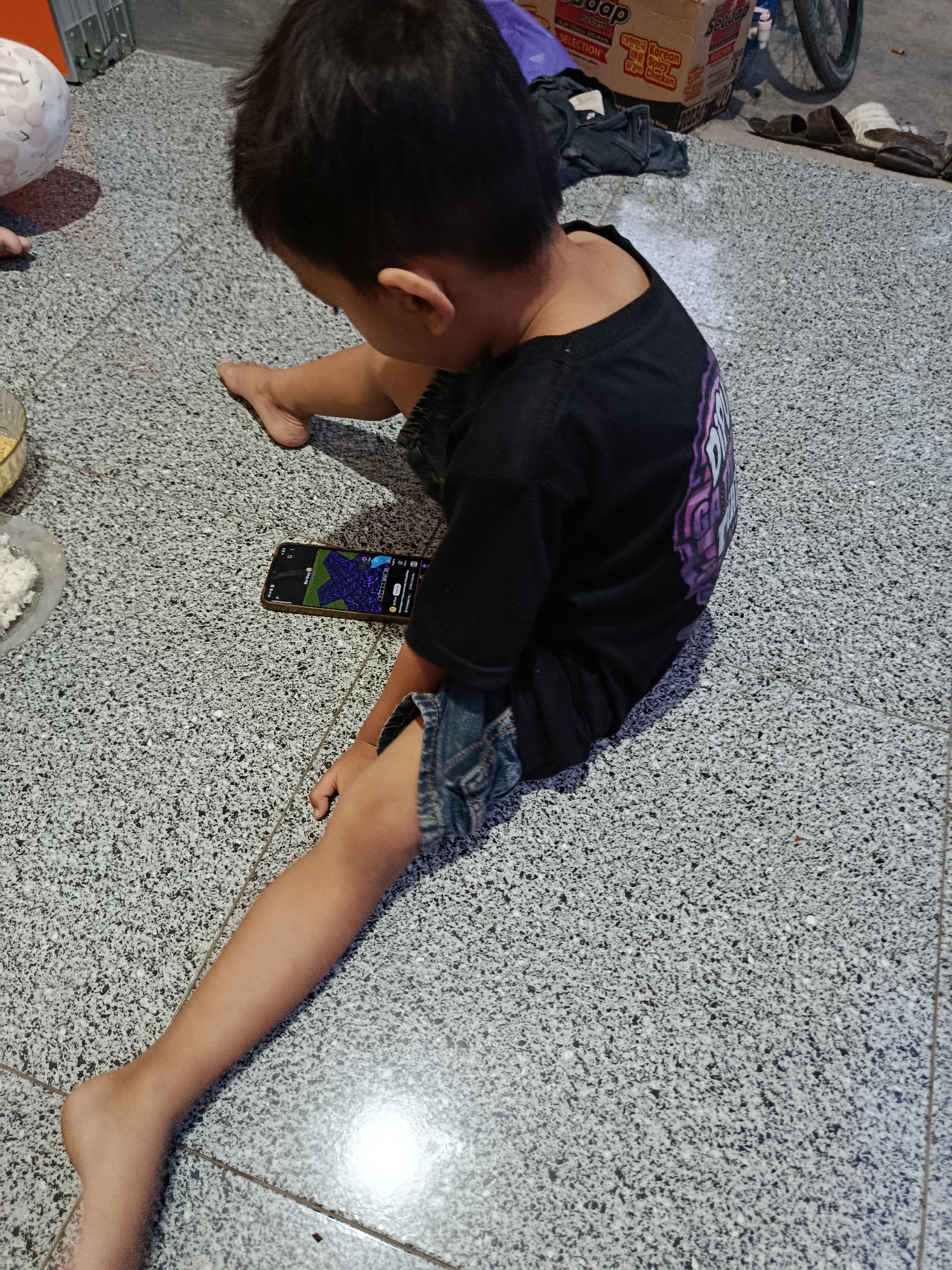 A young boy sits on a speckled grey floor, engrossed in a mobile phone. He is wearing a black t-shirt with a purple design and denim shorts. His left leg is extended in front of him, while his right leg is bent. A bowl of white rice and a glass bowl with yellow food are visible to the left of the boy. In the background, there are cardboard boxes, a bicycle wheel, and a pair of sandals. The scene appears to be an indoor setting, possibly a home in Sarwadadi, Indonesia.
