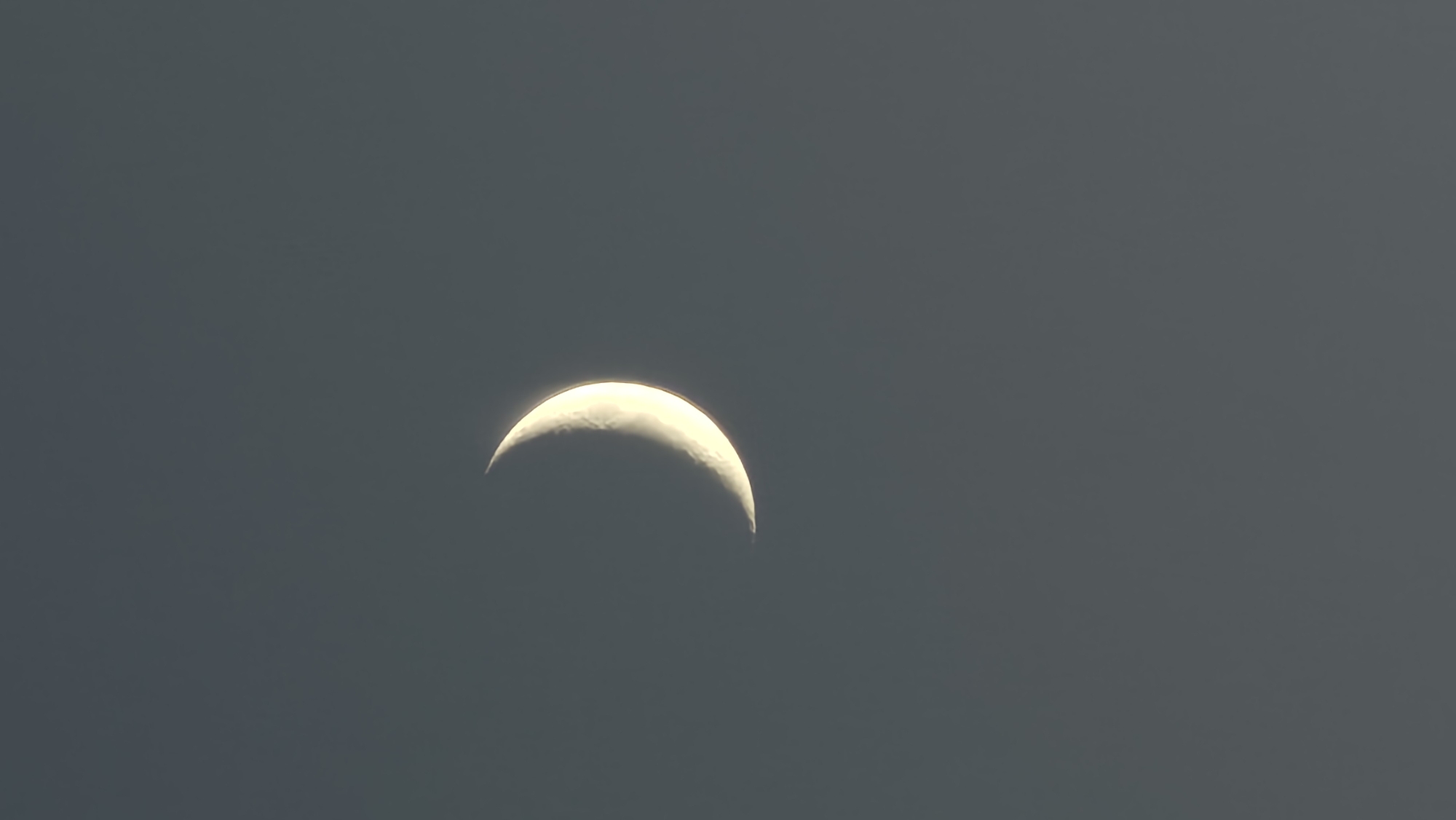 A crescent moon is visible in a dark, overcast sky. The moon is illuminated from the right side, casting a soft glow. The background is a uniform gray, suggesting a cloudy or twilight atmosphere. There are no discernible landmarks or environmental features indicating a specific location, including Yekaterinburg, Russia. The image is a close-up shot focused solely on the celestial body.
