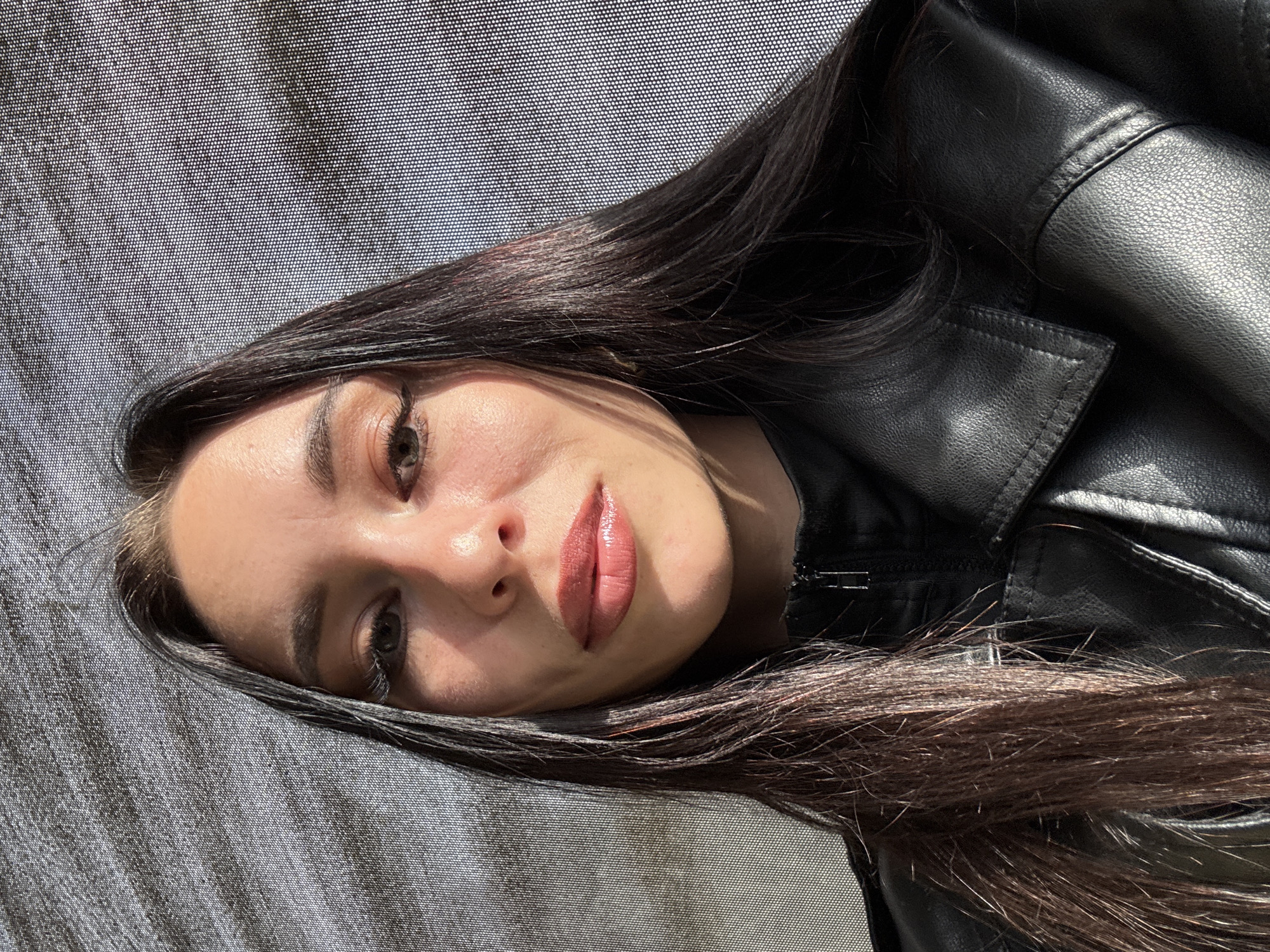 An eye-level close-up shows a young woman looking directly at the camera, positioned horizontally. Her face, seen from the mid-forehead to the chin, occupies the left side of the frame. She has dark, long hair that frames her face and extends into the bottom right. Her skin appears smooth, with natural light highlighting her forehead, nose, and lips, creating shadows on the side of her face. She has distinct eyebrows, dark eye makeup, and light-colored eyes. Her full lips are painted with a glossy, rosy-nude lipstick.
She is wearing a black leather jacket, visible on her right side, with visible stitching and a zipper detail near her neck. Her dark hair also rests against the jacket.
The background primarily consists of a textured, mesh-like fabric, appearing as a grid of small white dots against a darker gray/black material. This textured background suggests she might be lying down or reclining against a surface, possibly a car interior's ceiling or sunshade, or a textured wall. The bright, strong natural light, casting distinct shadows on her face, indicates it is daytime, likely sunny.
No discernible activity is taking place beyond posing for the photograph. Her expression is neutral and direct, suggesting an intentional gaze. There is no visible text in the image.