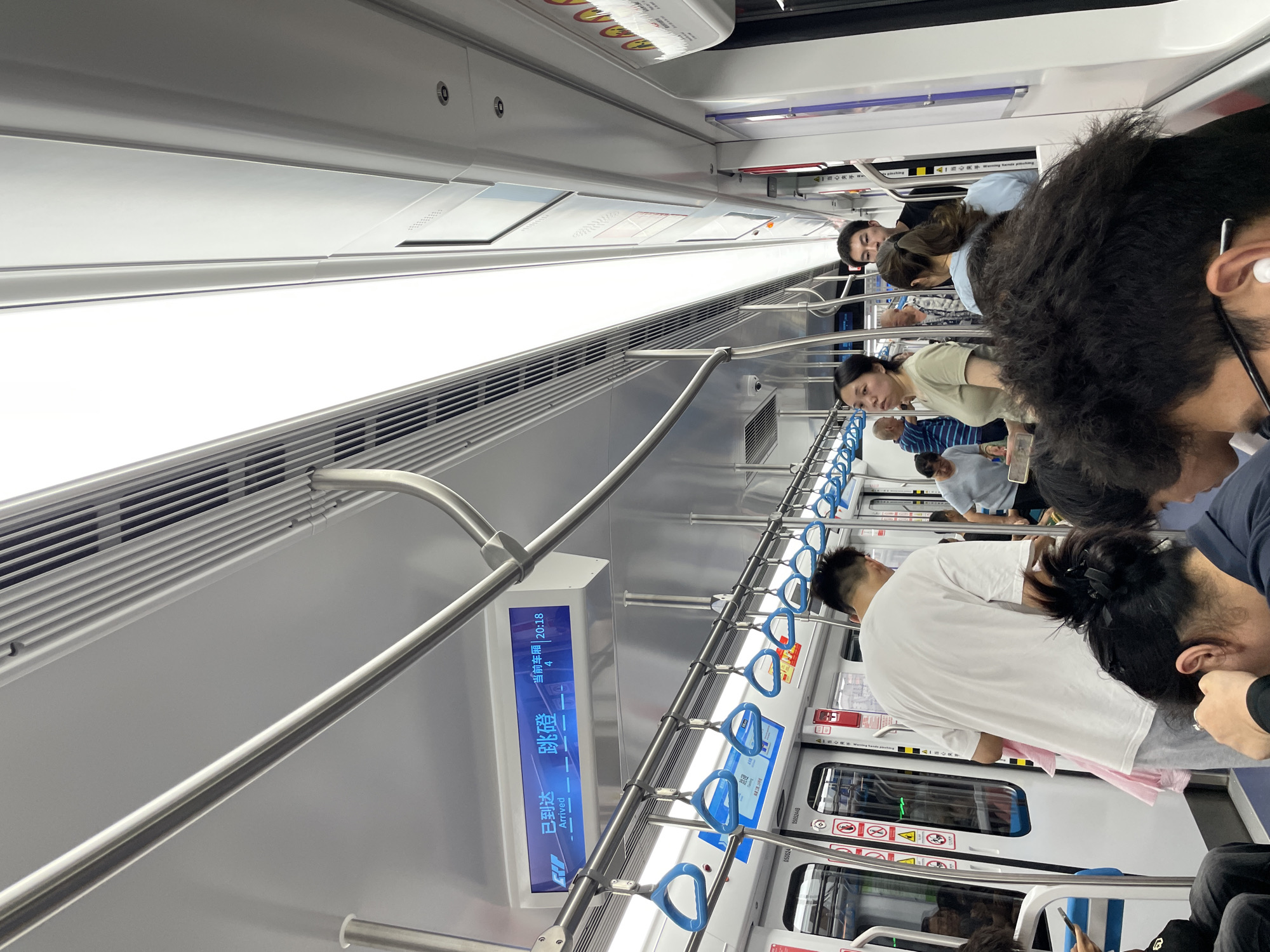 The image captures a scene inside a public transportation vehicle, possibly a train or subway, with several people standing and holding onto handrails or straps. The interior is modern, featuring light-colored panels, blue handholds, and a digital display indicating information such as "Arrived" and "20:18" alongside what appears to be Chinese characters. The environment is urban and functional, designed for transporting passengers efficiently.