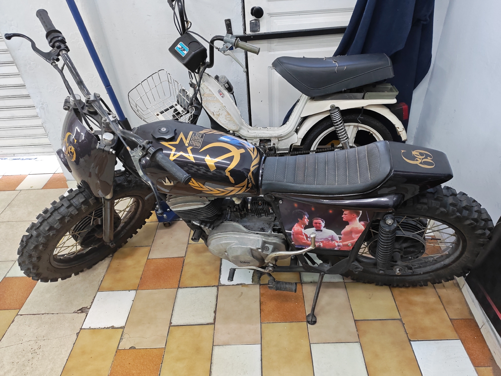 Two motorcycles are parked on a tiled floor with a grid pattern of beige, white, and brown.
The foreground motorcycle is black with gold accents and features a thick-treaded front tire. The side displays a boxing match graphic.
The background motorcycle is white, smaller, and has a wire basket attached to the handlebars. A blue cloth hangs nearby.
The location is an indoor space, likely a garage or shop. The exact location within Buenos Aires, Argentina, cannot be determined.