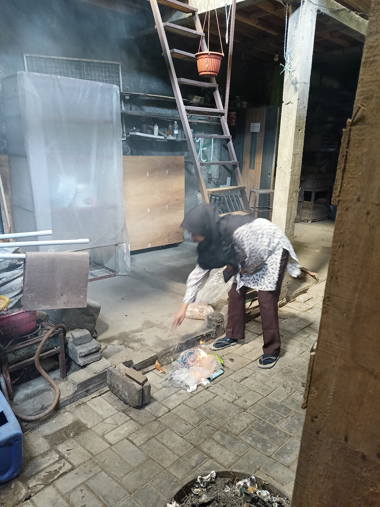 In Sukorini, Indonesia, a woman wearing a headscarf and patterned tunic is tending to a small, smoldering fire on a paved outdoor area. She is positioned near a pile of what appears to be burning trash, with plastic bags and other debris visible. The woman is reaching towards the fire with her right hand, possibly adding or adjusting the burning materials. The scene is dimly lit, with smoke rising from the fire and obscuring some of the background. The environment appears to be a backyard or an open-air workspace, with various objects scattered around, including a metal ladder, shelves with items, and construction materials. The ground is paved with bricks.