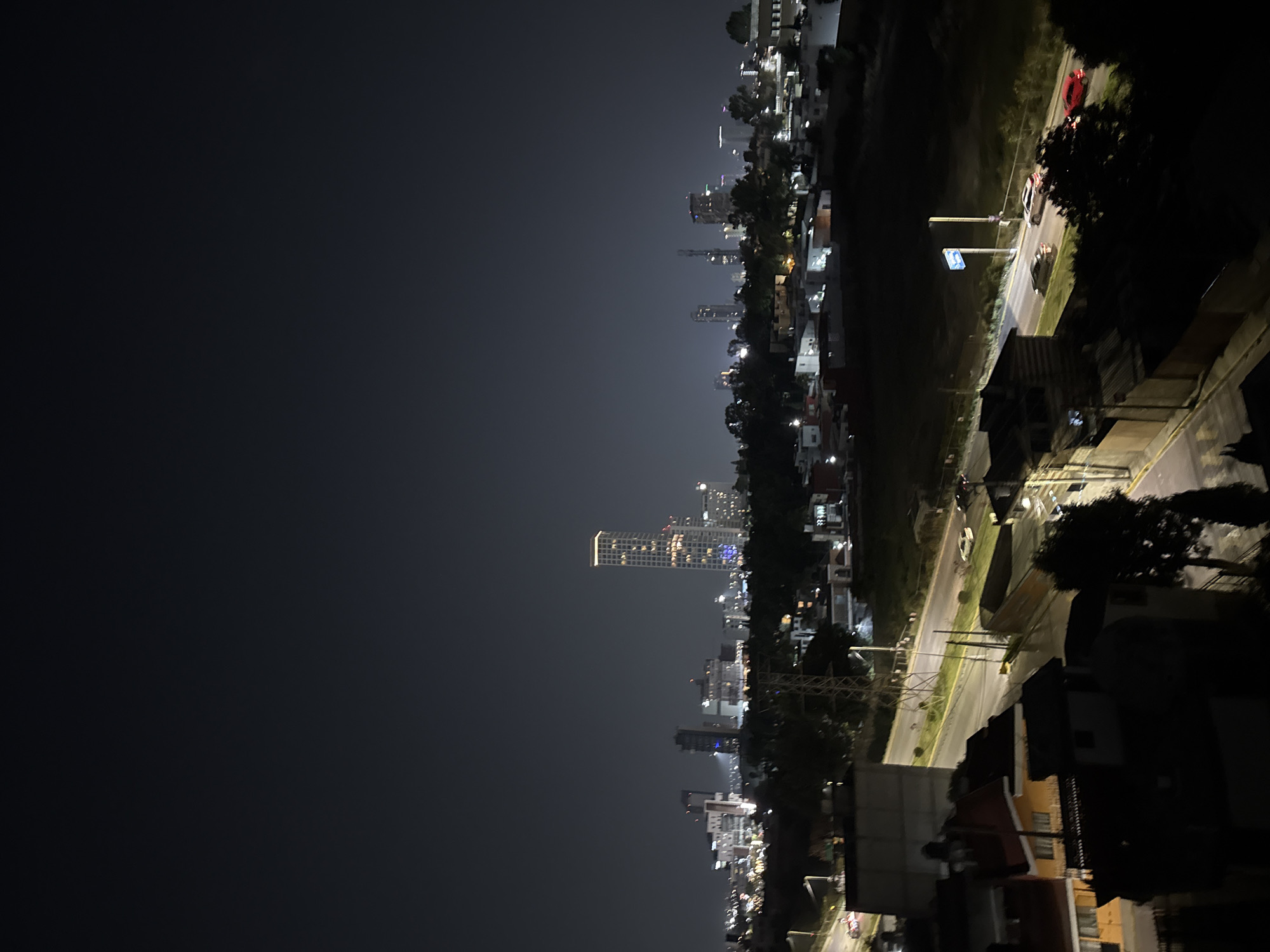 The image depicts a nighttime view of San Bernardino Tlaxcalancingo, Mexico. A major thoroughfare illuminated by streetlights is visible, with several vehicles moving along the road. Buildings line the right side of the frame, ranging from residential structures to taller buildings, with glimpses of urban greenery interspersed. In the distance, high-rise buildings of a larger city are discernable against the night sky, lit by the city's night lights. The sky is dark, transitioning from a lighter gray near the horizon to a deep black. The overall setting suggests an urban or suburban environment with a well-lit road network.