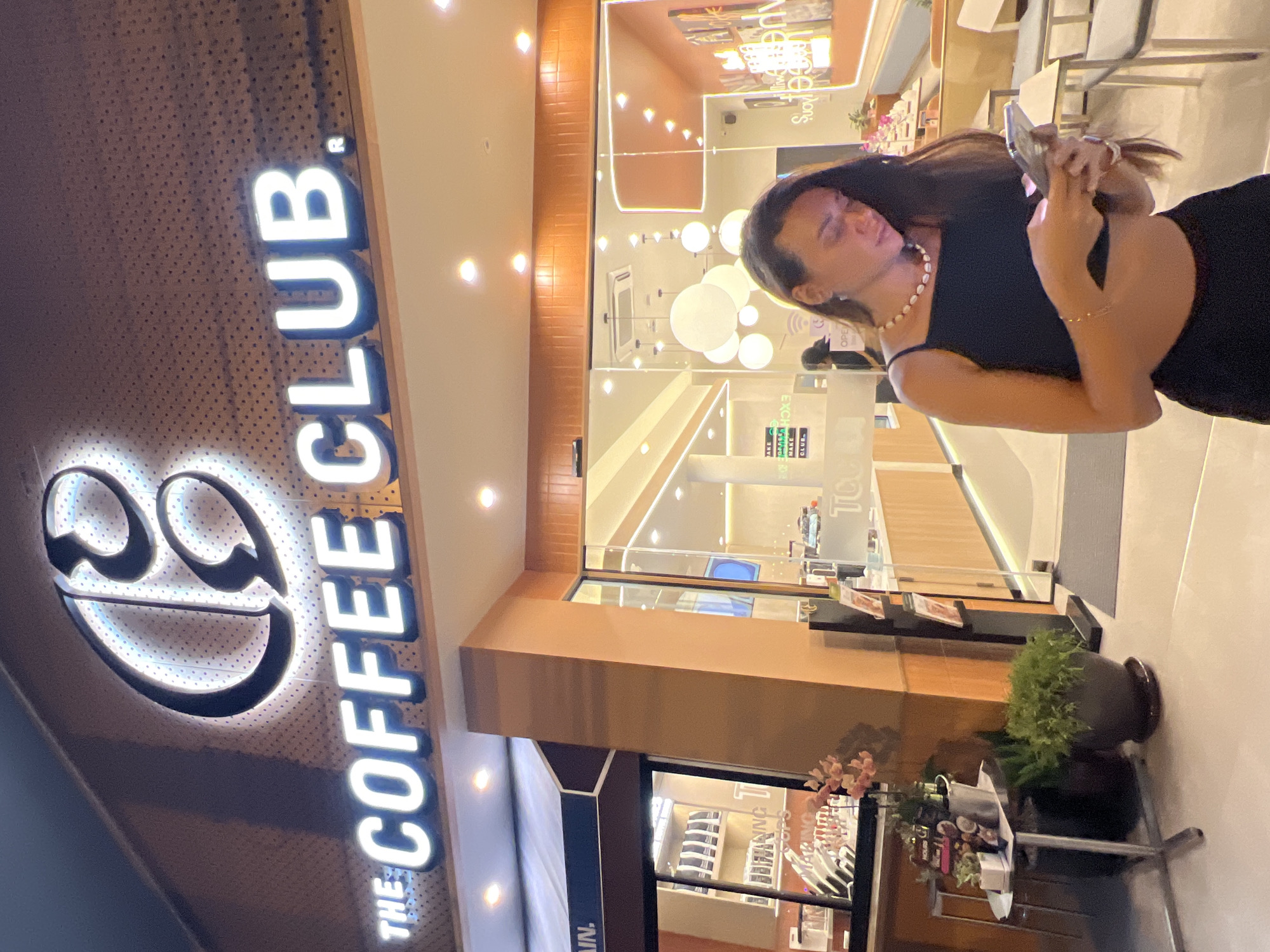 A woman with long brown hair is standing outside "The Coffee Club" store in Baan Bo Phut, Thailand. She is wearing a black dress and holding a smartphone. The store has a lit logo and large glass windows. There are potted plants near the entrance.