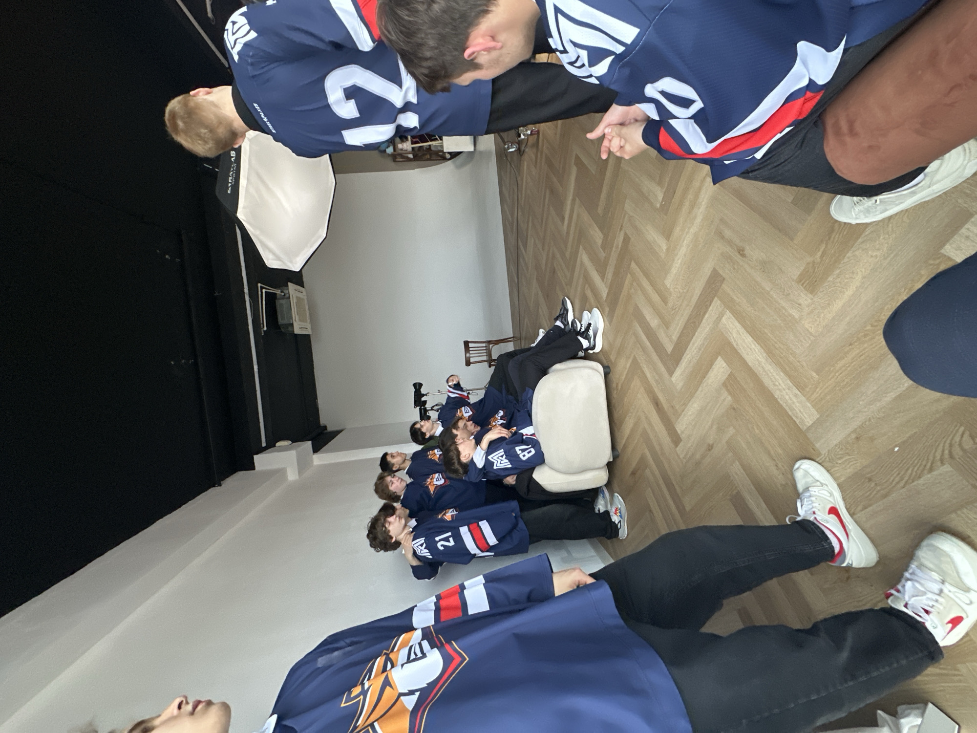 A group of people wearing hockey jerseys are resting on an upholstered bench in a room with a herringbone wood floor. The room has white walls and a black backdrop. There is lighting equipment present. The city and location are unknown.