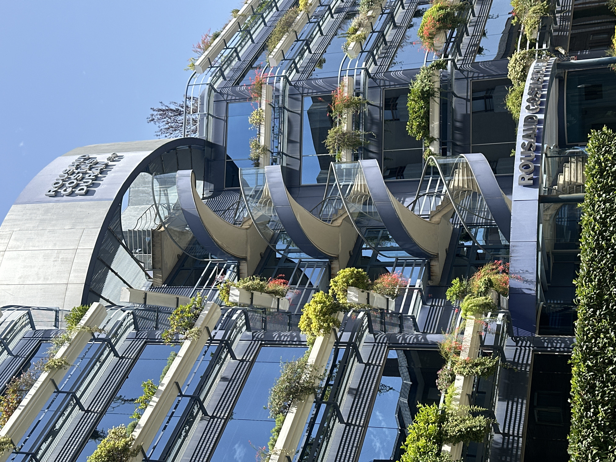 This is a daytime, eye-level, medium shot of the ROUSAUD COSTA DURAN building in Barcelona, Spain. The building's facade is a mix of glass and metal, with multiple levels of balconies adorned with planters overflowing with lush greenery and colorful flowers. A prominent blue spiral staircase winds its way up the side of the building, providing access to different floors. The sky above is a clear blue. The entrance to the building is visible at the bottom, with the name "ROUSAUD COSTA DURAN" displayed in metallic lettering above it. A dense hedge runs along the bottom of the image, framing the entrance and the lower part of the building. The scene conveys a modern architectural style integrated with natural elements.