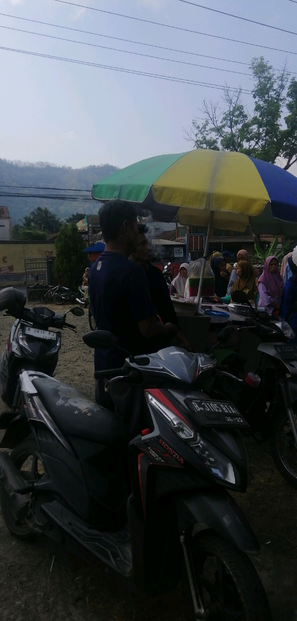 The image depicts a street scene in Windunegara, Indonesia, under a bright, cloudy sky. In the foreground, two motorcycles are parked. The one on the left is dark with a white and black seat, and its license plate reads "R 2810 PJ". The motorcycle on the right is primarily black and red, with the brand "HONDA" clearly visible. Its license plate is "B-3105 BIA". Behind the motorcycles, a colorful umbrella, segmented in yellow, green, and blue, provides shade for a small roadside stall. Several people are gathered around the stall. A man in a dark blue t-shirt is in the left-center of the frame, looking away from the camera and smoking. To his right, another man is partially visible. Further to the right, several women, some wearing headscarves (hijabs) in various colors like purple, brown, and white, are interacting at the stall. One woman in a purple hijab is holding a clear plastic bag containing a green and red layered liquid. A bag of what appears to be packaged food or drinks is also visible on the counter. In the background, there are buildings, some trees, and a distant, hazy hill. Power lines stretch across the top of the frame. The ground appears to be unpaved, likely dirt or gravel. The overall impression is a typical street-side vending scene in a somewhat rural or suburban area.