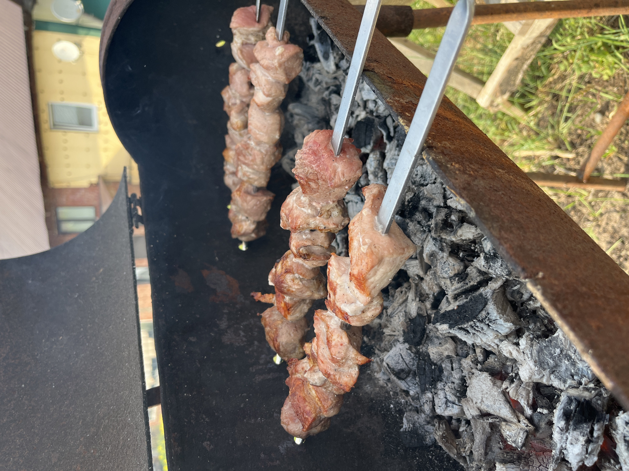 This is a medium shot of raw meat skewers cooking over charcoal in a barbecue grill. The meat appears to be pork, cut into cubes and threaded onto metal skewers. Some of the meat is already showing signs of browning and charring from the heat. The grill is made of dark metal and is filled with glowing red embers and ash. In the background, a yellow house with a brown roof is visible, suggesting a residential area. The overall impression is that of an outdoor cooking scene, likely in a backyard or park. The context of Chaikovsky, Russia, implies this is a common summer activity in that region.